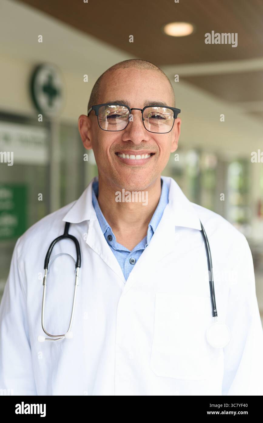 Reifer männlicher Arzt trägt Labormantel und Brille, der selbstbewusst vor einer kleinen Gesundheitsklinik steht. Medizinisches Fachkonzept für den Außenbereich. Stockfoto