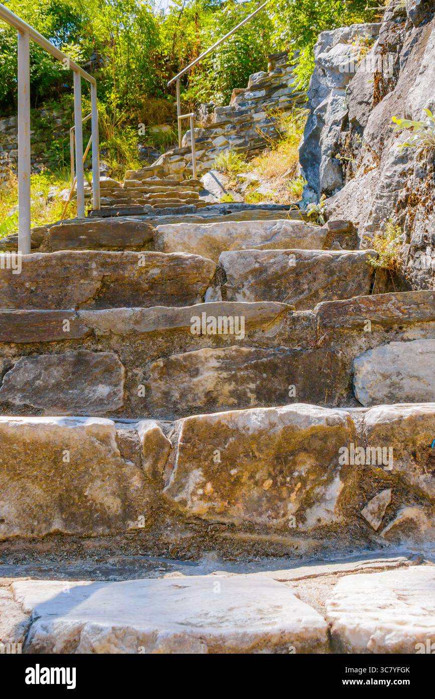 Grob gehauene Steinstufen führen sich an einem steilen Hügel entlang, flankiert von üppigem Grün und begleitet von einem stabilen Metallgeländer. Stockfoto