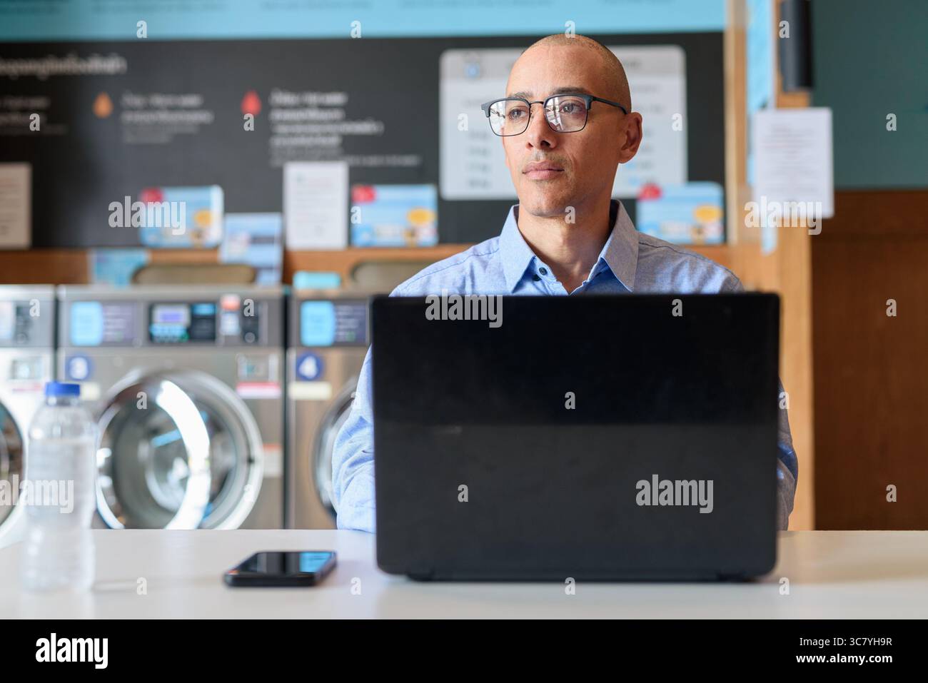 Glatzköpfiger männlicher Profi in blauem Button-Down-Hemd, der in der Wäscherei am Laptop arbeitet. Er setzt Technologie im Alltag ein. Echte Arbeit Stockfoto