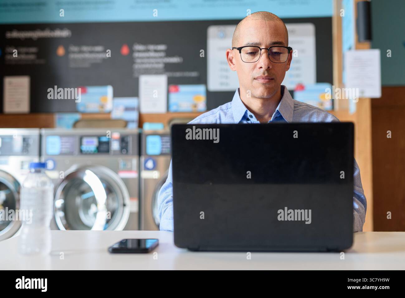 Glatzköpfiger männlicher Profi in blauem Button-Down-Hemd, der in der Wäscherei am Laptop arbeitet. Er setzt Technologie im Alltag ein. Echte Arbeit Stockfoto