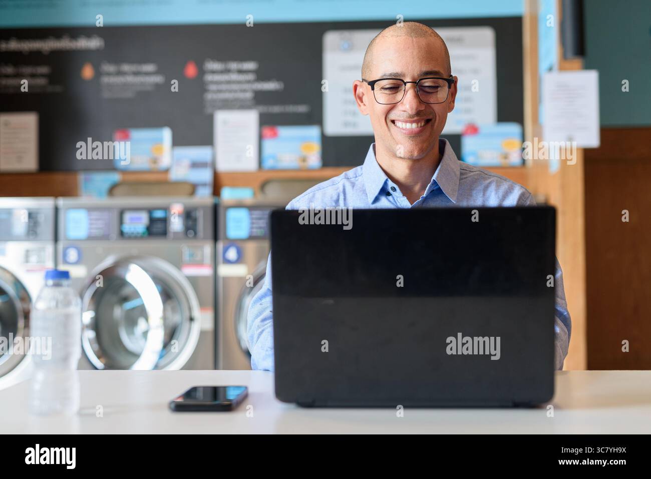 Glatzköpfiger männlicher Profi in blauem Button-Down-Hemd, der in der Wäscherei am Laptop arbeitet. Er setzt Technologie im Alltag ein. Echte Arbeit Stockfoto