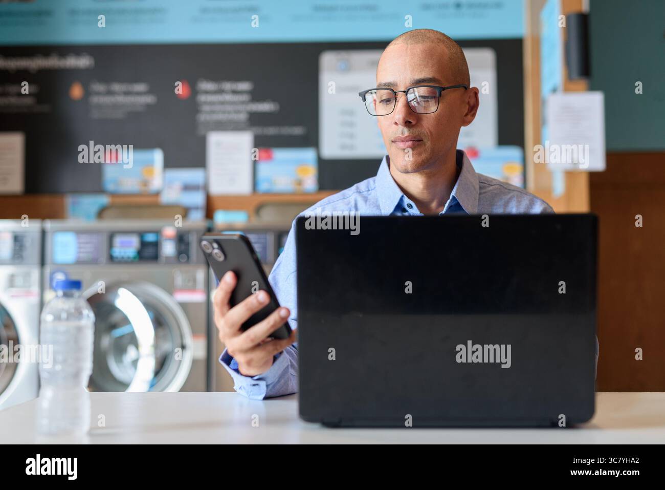Glatzköpfiger männlicher Profi in blauem Button-Down-Hemd, der in der Wäscherei am Laptop arbeitet. Er setzt Technologie im Alltag ein. Echte Arbeit Stockfoto