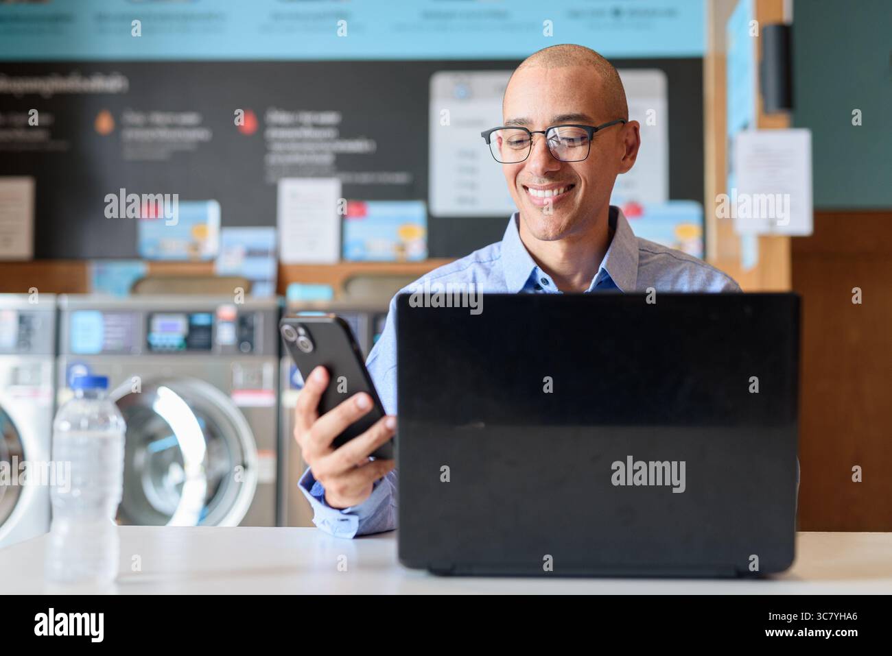 Glatzköpfiger männlicher Profi in blauem Button-Down-Hemd, der in der Wäscherei am Laptop arbeitet. Er setzt Technologie im Alltag ein. Echte Arbeit Stockfoto