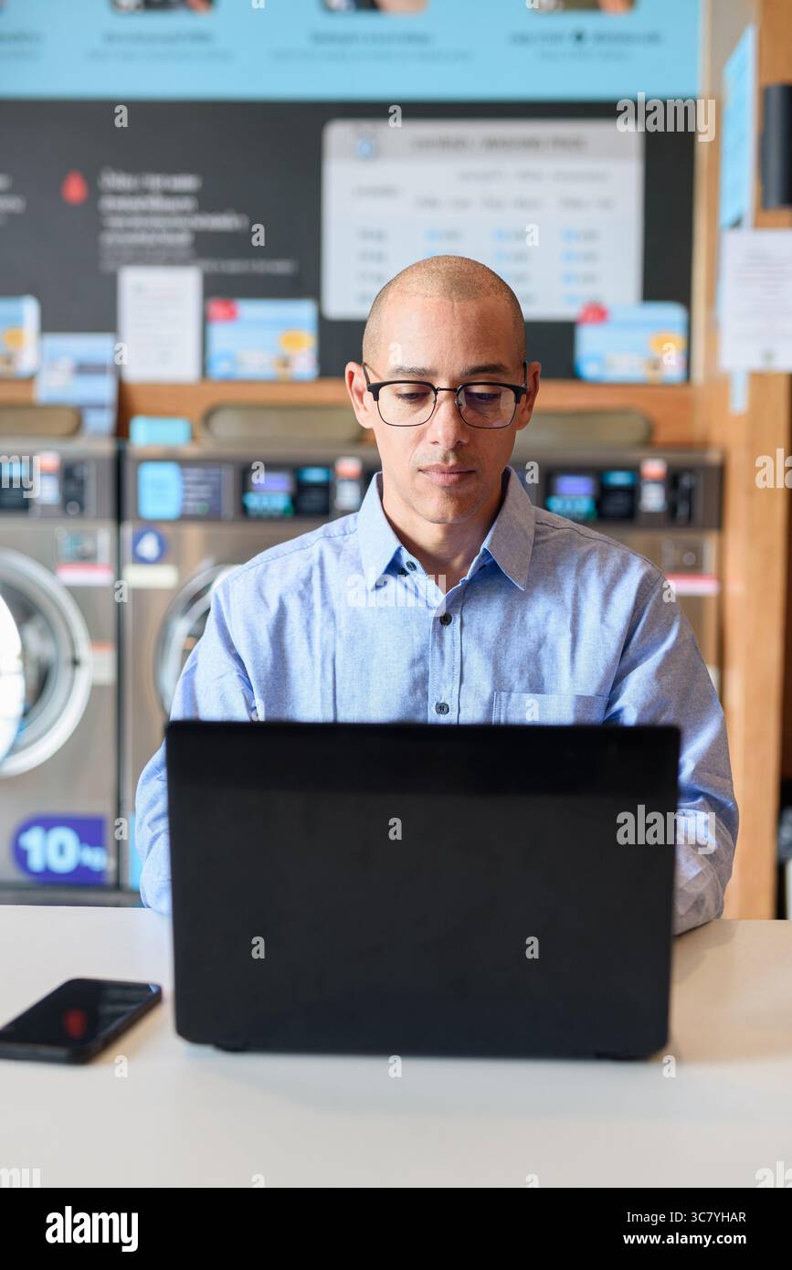 Glatzköpfiger männlicher Profi in blauem Button-Down-Hemd, der in der Wäscherei am Laptop arbeitet. Er setzt Technologie im Alltag ein. Echte Arbeit Stockfoto
