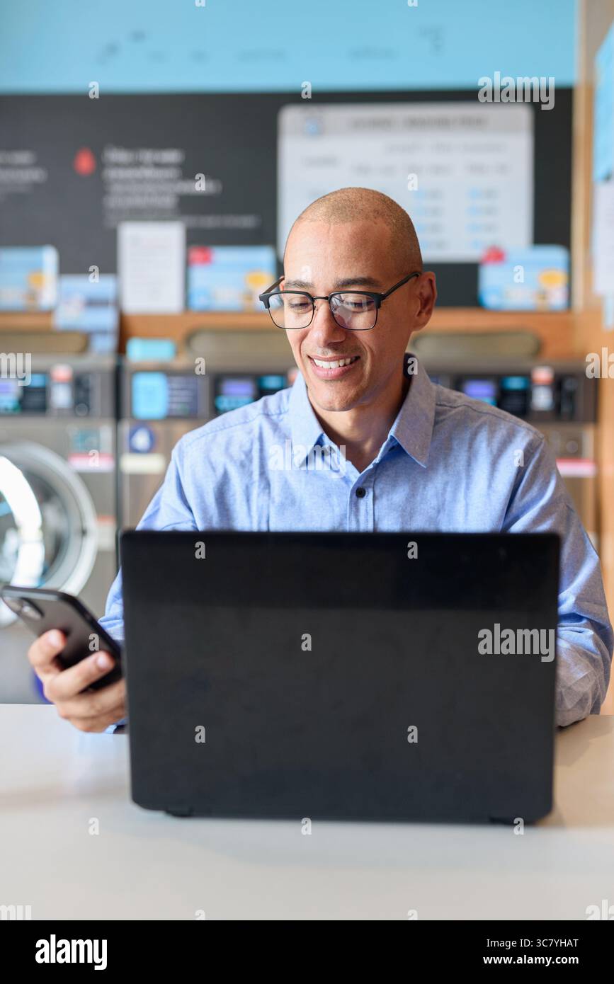 Glatzköpfiger männlicher Profi in blauem Button-Down-Hemd, der in der Wäscherei am Laptop arbeitet. Er setzt Technologie im Alltag ein. Echte Arbeit Stockfoto