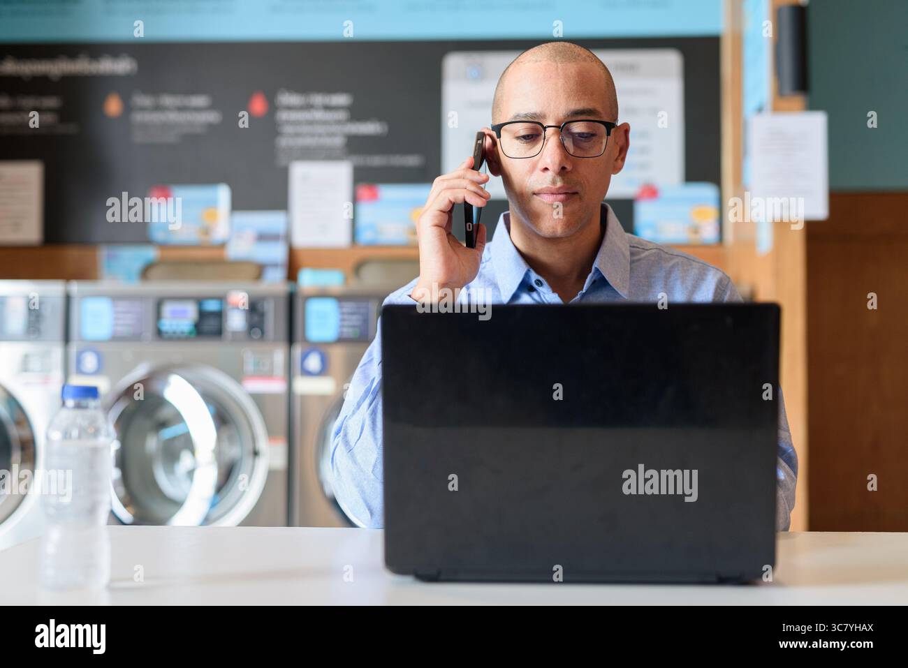 Glatzköpfiger männlicher Profi in blauem Button-Down-Hemd, der in der Wäscherei am Laptop arbeitet. Er setzt Technologie im Alltag ein. Echte Arbeit Stockfoto