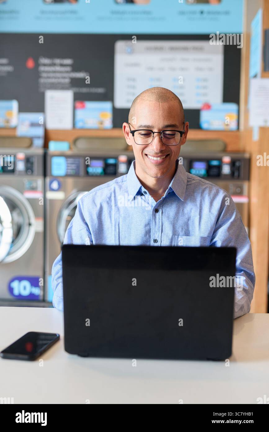Glatzköpfiger männlicher Profi in blauem Button-Down-Hemd, der in der Wäscherei am Laptop arbeitet. Er setzt Technologie im Alltag ein. Echte Arbeit Stockfoto