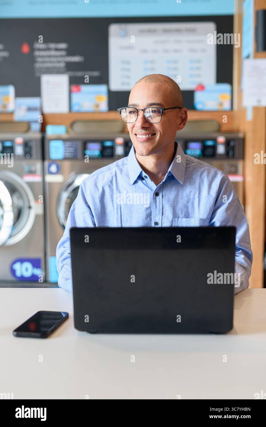 Glatzköpfiger männlicher Profi in blauem Button-Down-Hemd, der in der Wäscherei am Laptop arbeitet. Er setzt Technologie im Alltag ein. Echte Arbeit Stockfoto