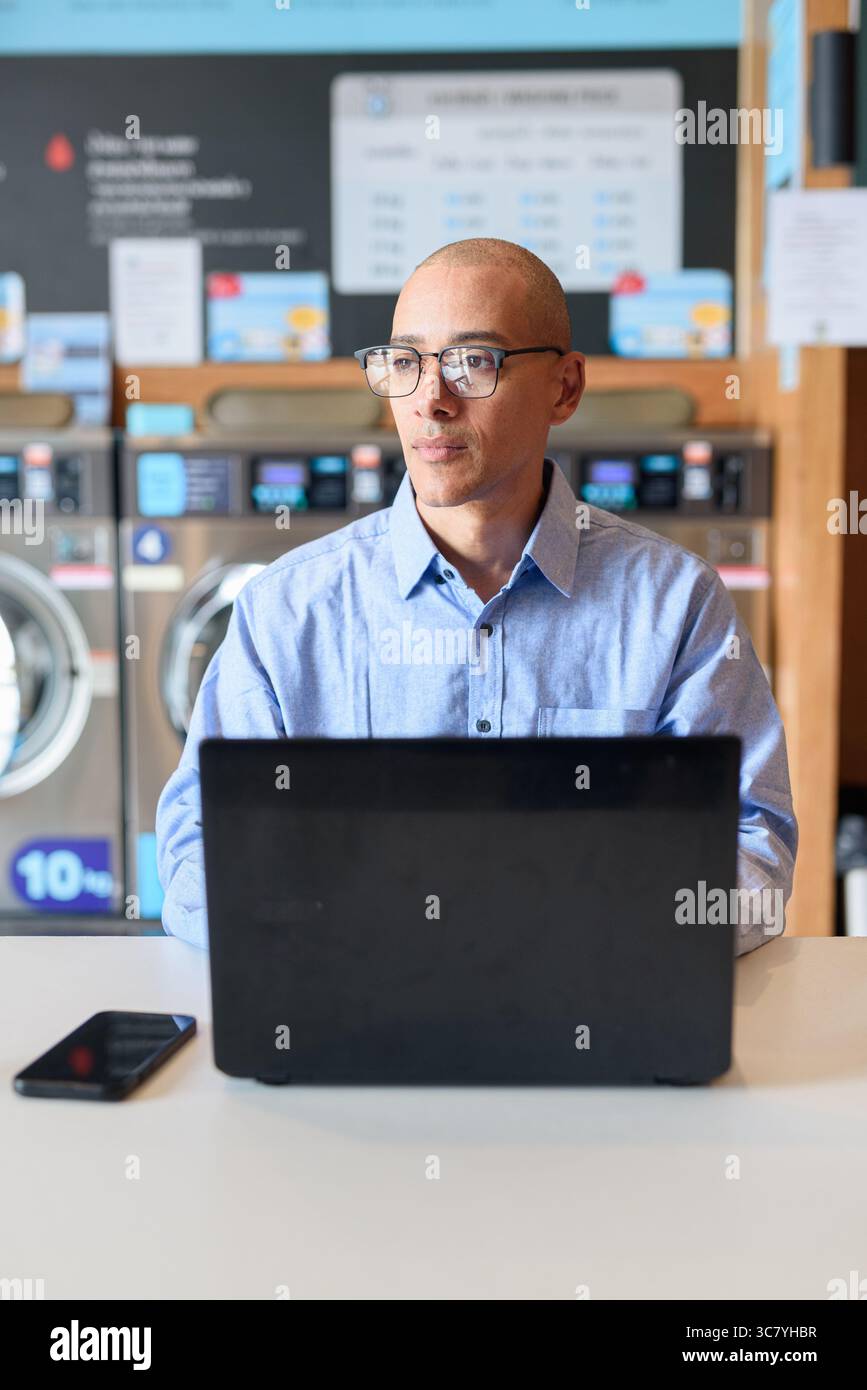 Glatzköpfiger männlicher Profi in blauem Button-Down-Hemd, der in der Wäscherei am Laptop arbeitet. Er setzt Technologie im Alltag ein. Echte Arbeit Stockfoto