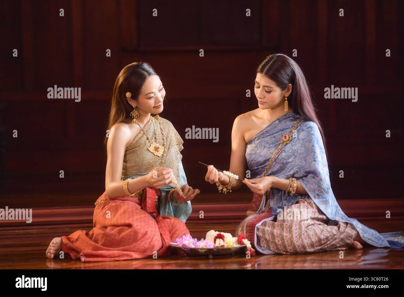 Zwei junge Frauen in traditioneller thailändischer Kleidung sitzen nebeneinander auf dem Boden und machen Blumengirlanden (Phuang Malai) Stockfoto