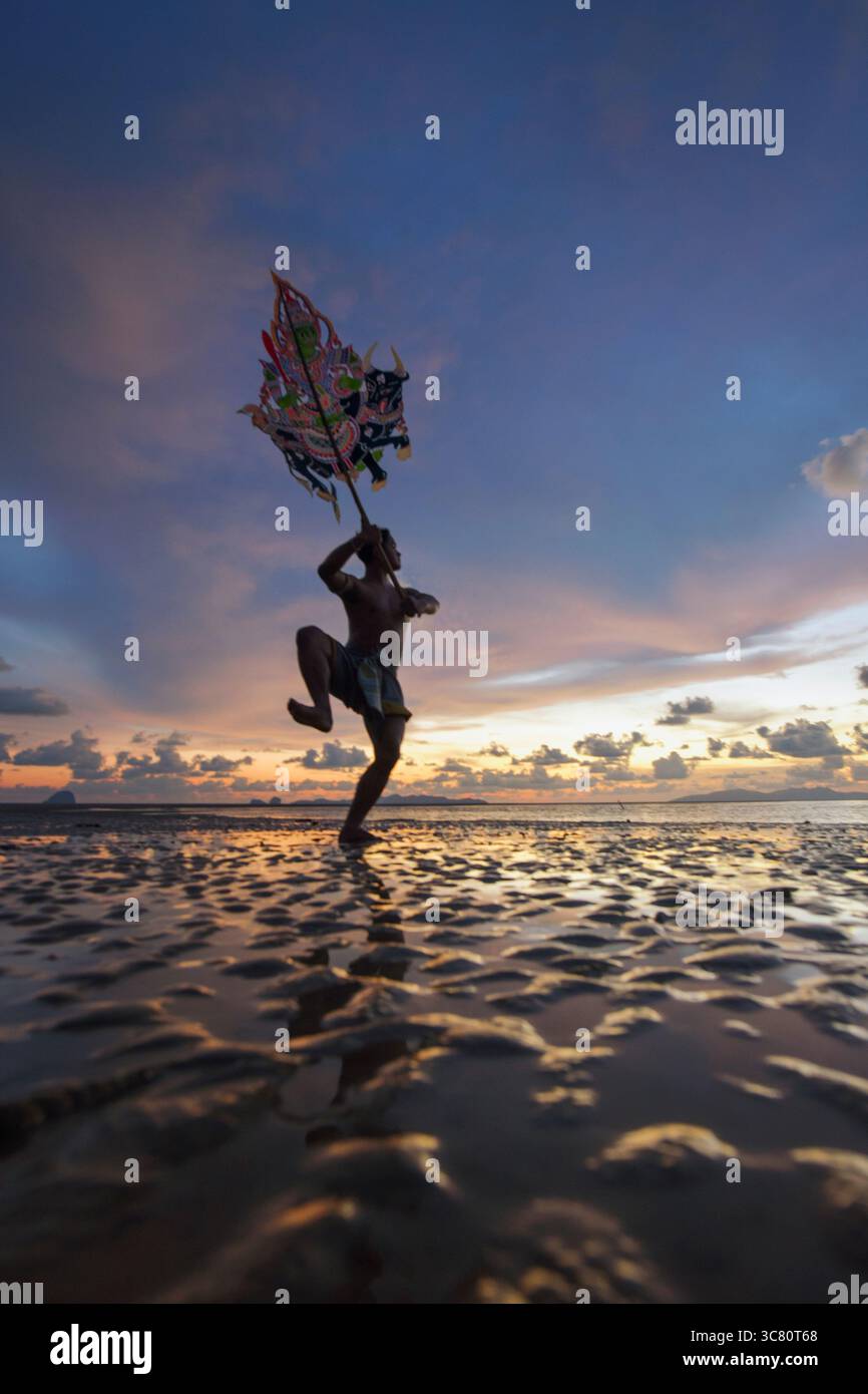 Silhouette eines Mannes, der die traditionelle Thai Shadow Puppet Show (Nang Talung) an einem Strand bei Sonnenaufgang in Thailand aufführt Stockfoto