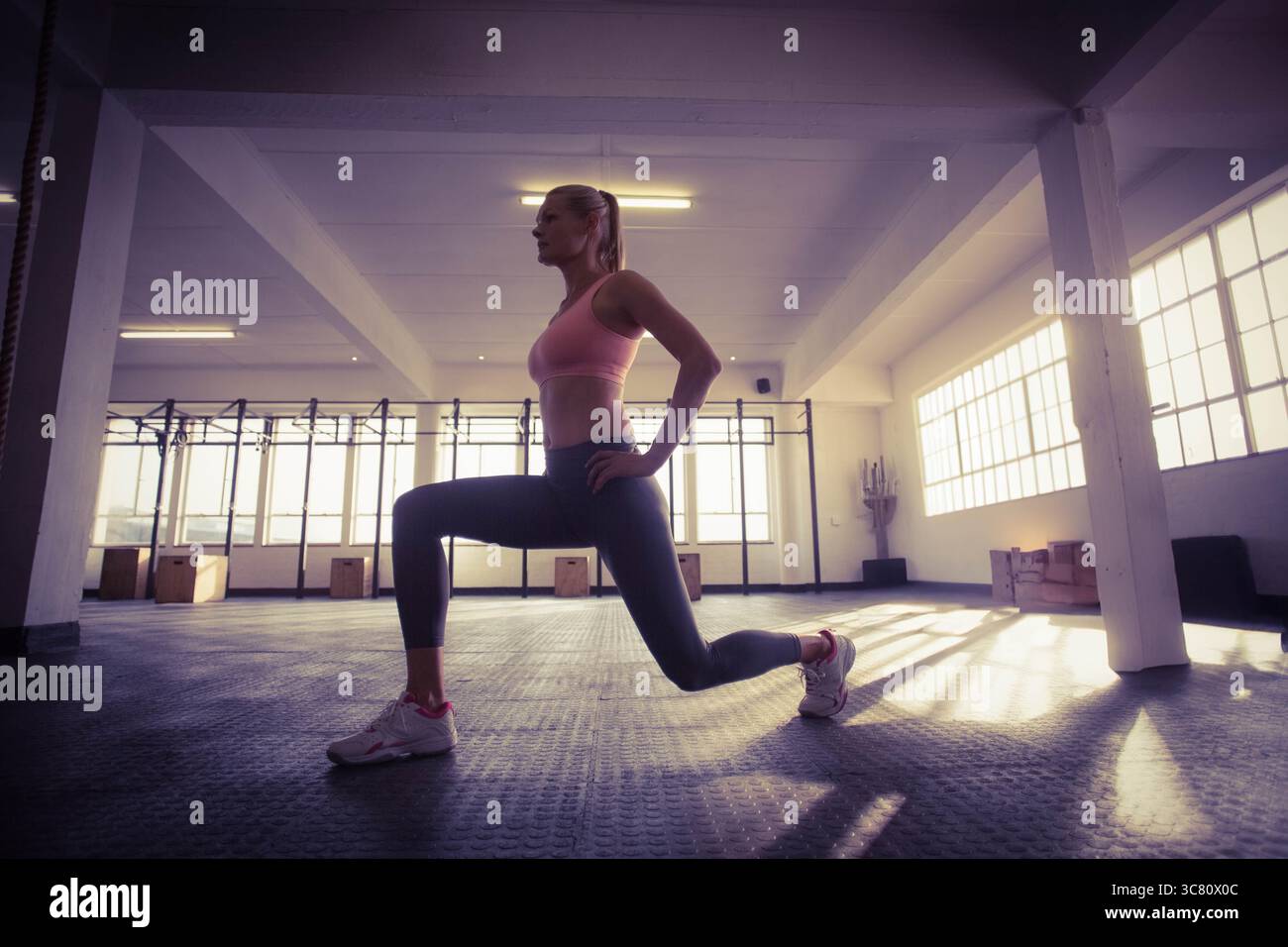 Eine Frau, die einen Ausfallschritt auf einem strukturierten Gummi-Fitnessfußboden mit einem Klimmfahrzeug, einem Seil, Plyoboxen in der Nähe von Fenstern macht Stockfoto