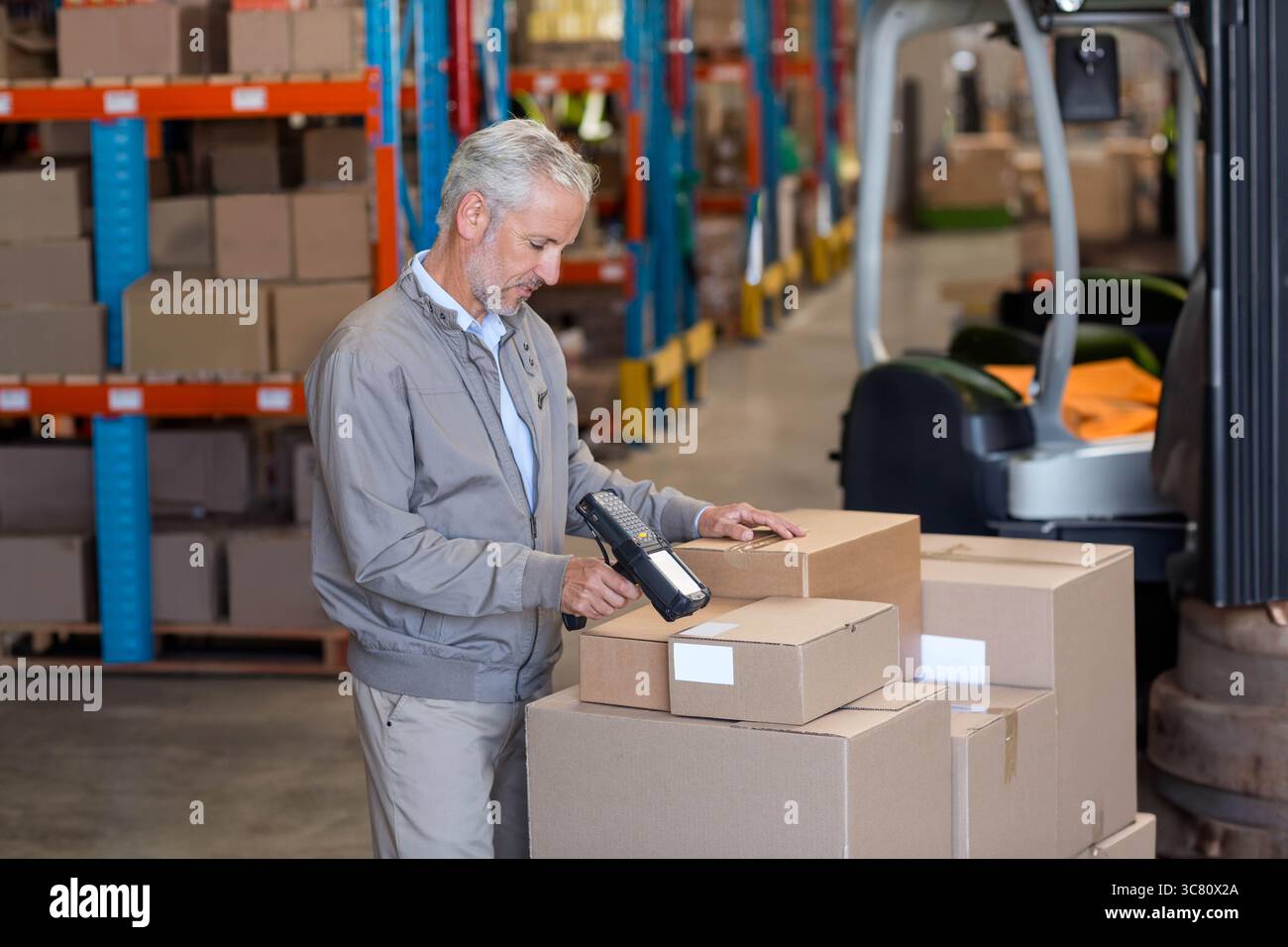 Lagerarbeiter scannt Kartons mit Strichcode-Scanner zwischen Regalen in der Nähe eines geparkten Gabelstaplers Stockfoto