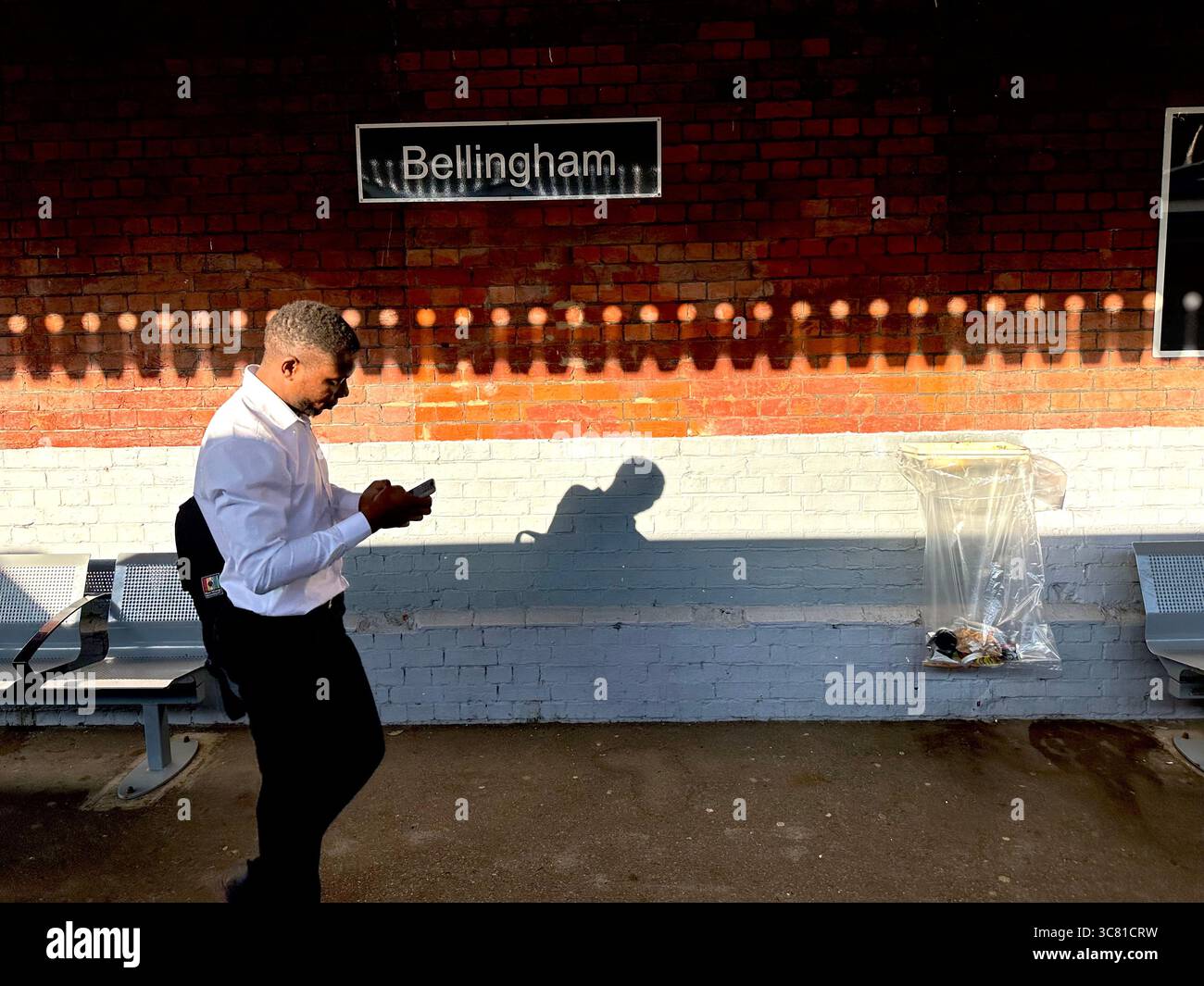Ein Mann mit einem Rucksack, der sein Smartphone ansieht und SMS schreibt, während er am Bahnhof Bellingham läuft - Smartphone-aufgenommenes Stockfoto