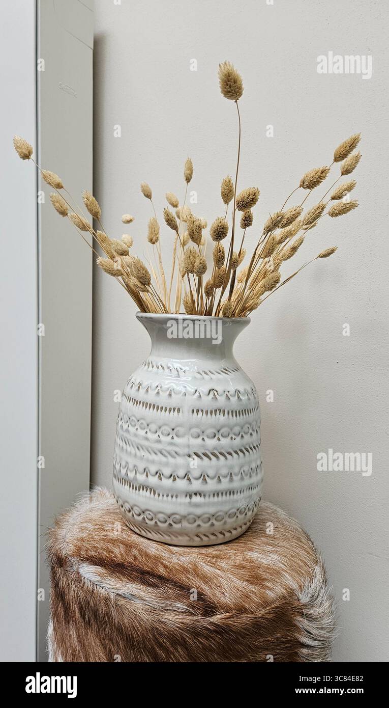 Dekorative Keramikvase mit geätzten Mustern, gefüllt mit getrockneten Gräsern, platziert auf einem Kunstfellhocker. - Smartphone-aufgenommenes Stockfoto