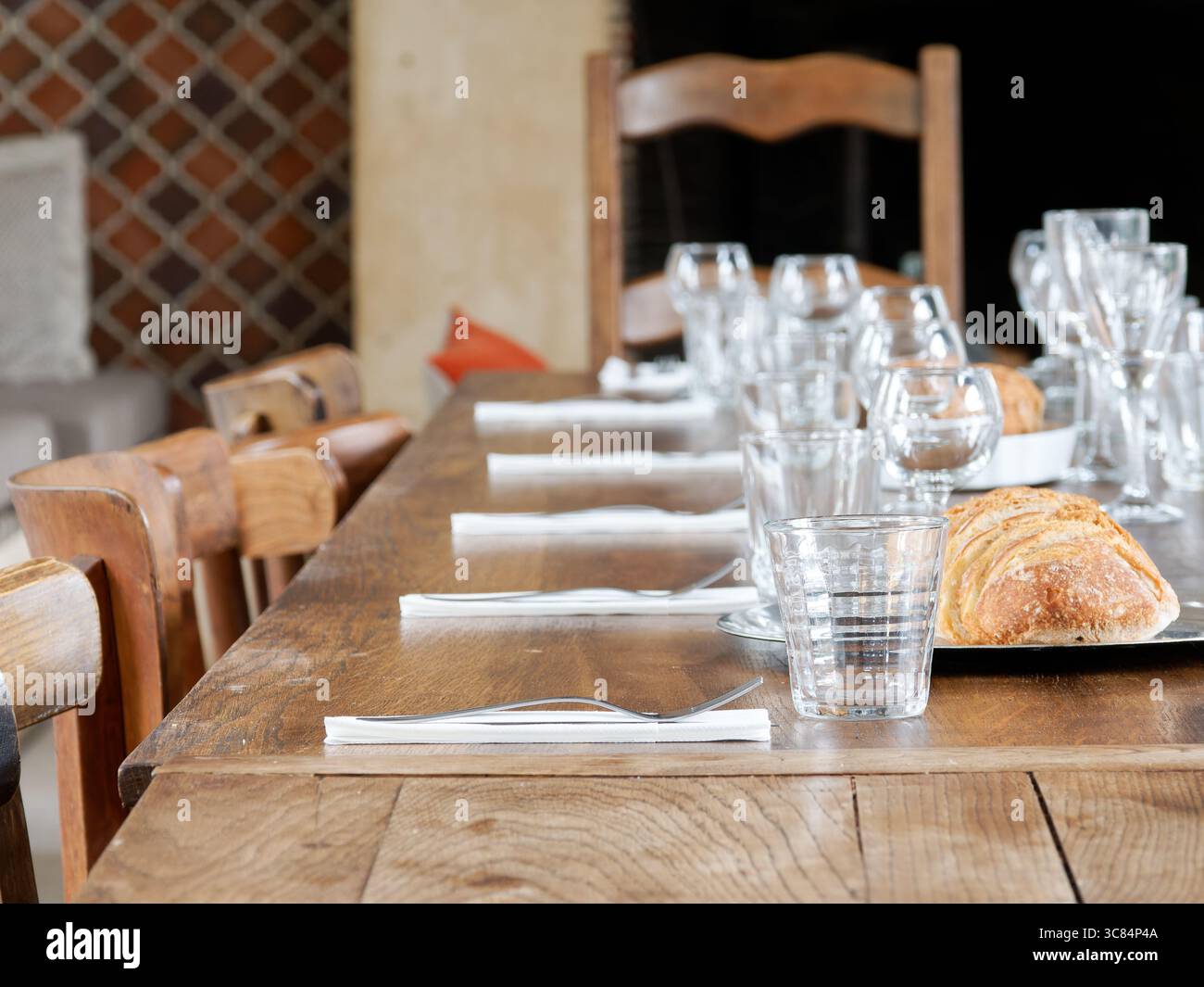 Ein langer rustikaler Holztisch für ein festliches Essen im Innenbereich, mit klaren Gläsern, weißen Servietten und einem Laib handwerklich geschnittenem Brot. Stockfoto
