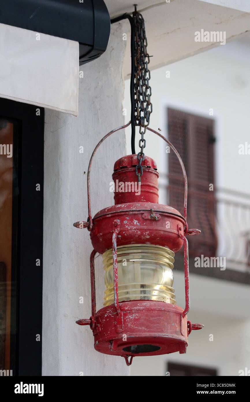 Eine alte rote Seefahrerlaterne aus Metall und Glas hängt an einer Kette unter einer Veranda und zeigt Anzeichen von Rost und Witterung von draußen Stockfoto