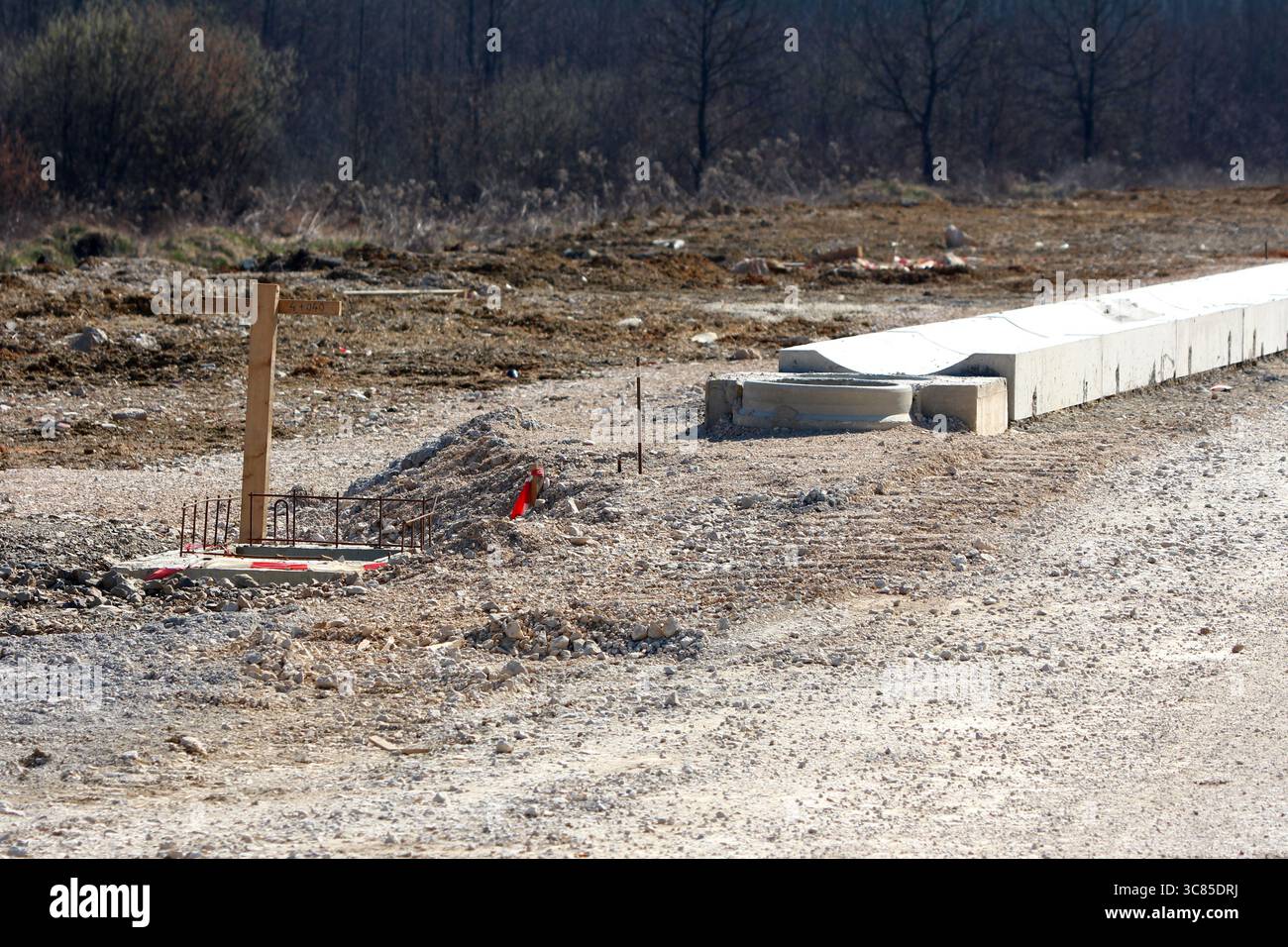 Auf der unbefestigten Straße befindet sich ein hölzerner Übersichtsmarker, der in Beton, freiliegende Bewehrungsstäbe und Betonfertigteile eingebettet ist, um die frühen Phasen der Fahrbahn zu veranschaulichen Stockfoto