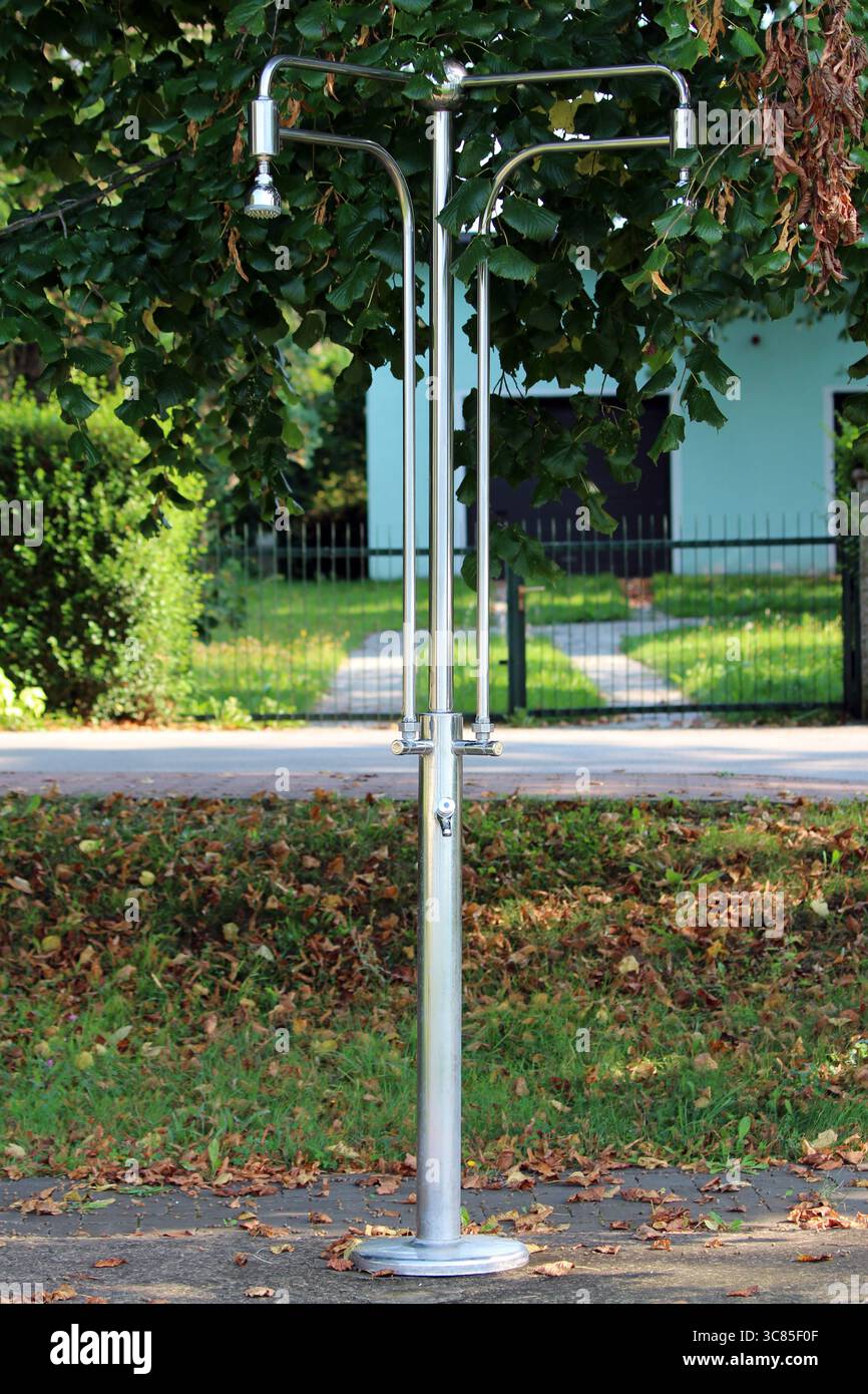 Zwei Edelstahl-Duschköpfe und -Armaturen stehen nebeneinander auf einer zentralen Metallsäule, umgeben von Grün und einem ruhigen Wohnhaus Stockfoto