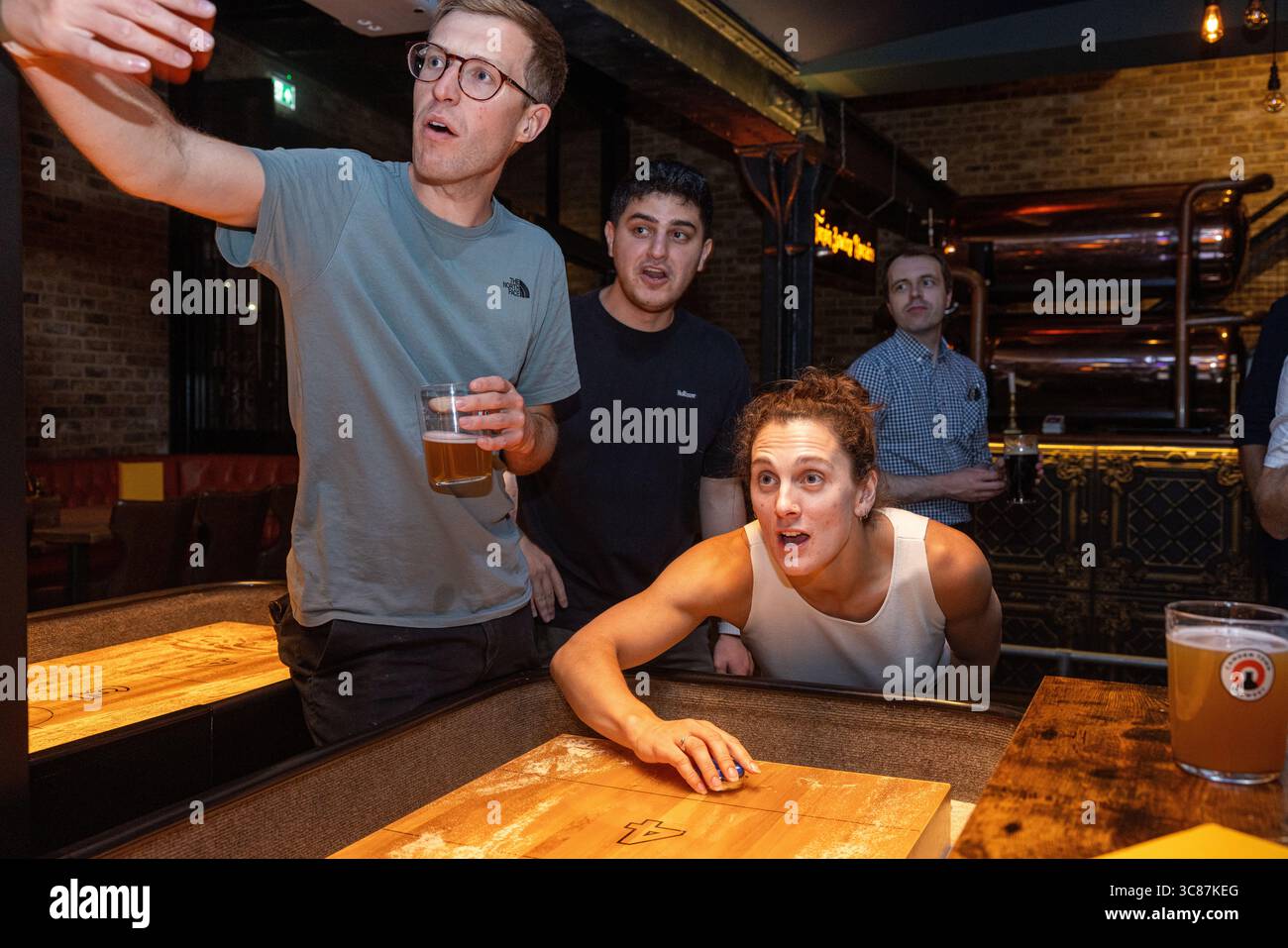 Junge Leute spielen Shuffleboard und machen gemeinsam ein Selfie in einer lebhaften Bar, um einen unterhaltsamen und geselligen Moment während eines Abends festzuhalten. Stockfoto