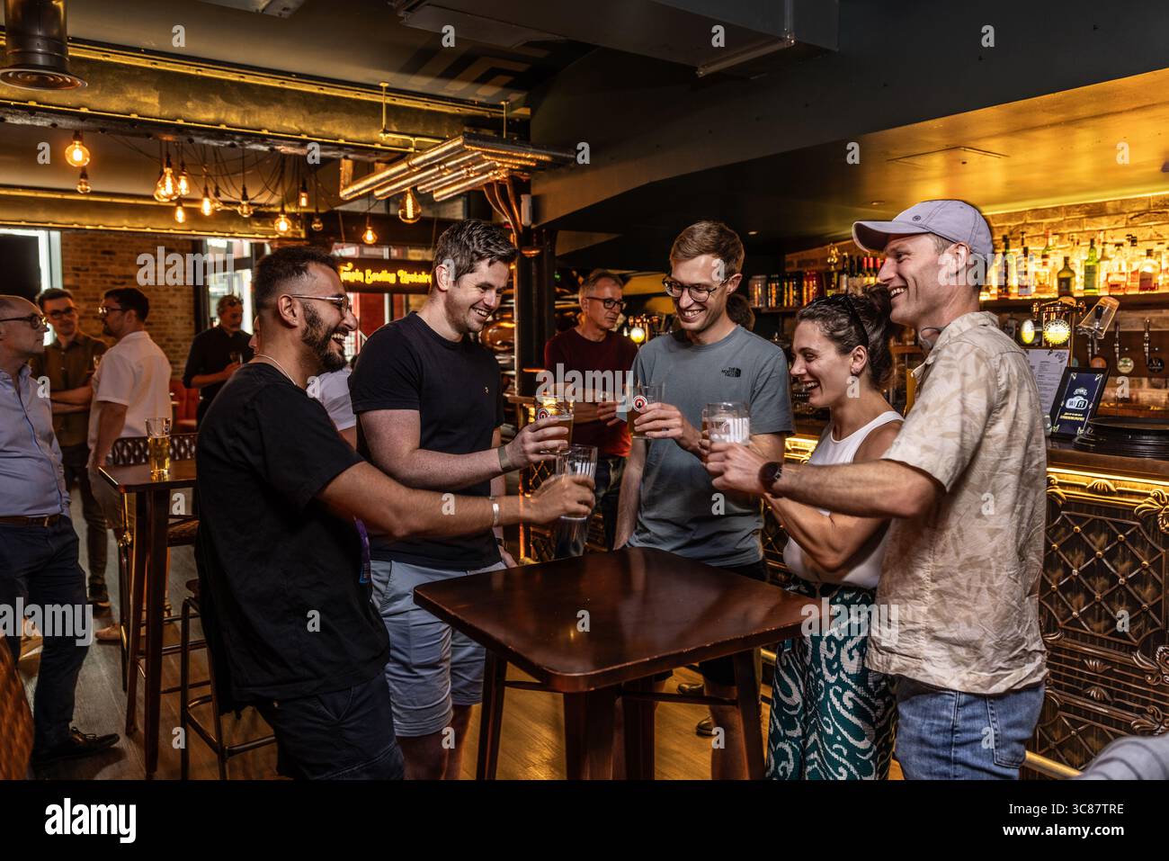 Gruppe junger Profis, die in einer lebhaften Bar Biergläser anklitzen, After-Work-Drinks und Teamarbeit in zwangloser Atmosphäre genießen. Stockfoto