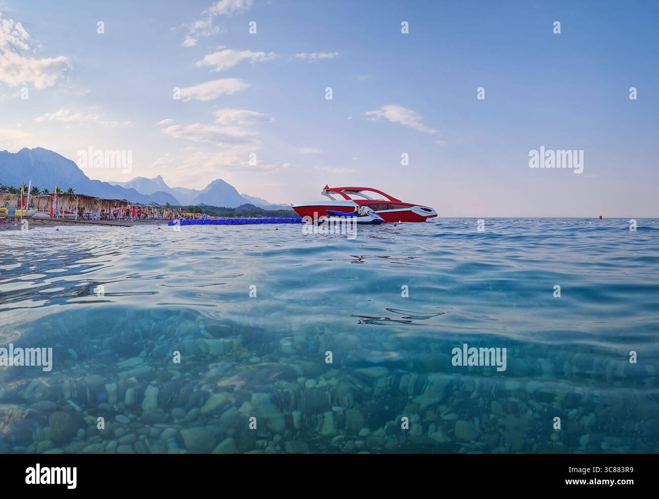 Panoramaaufnahme des Kemer Strandes mit klarem türkisfarbenem Wasser, das Kieselsteine darunter freilegt. Schnellboote ankerten nahe der Küste, und wundervolle Taurusberge i Stockfoto
