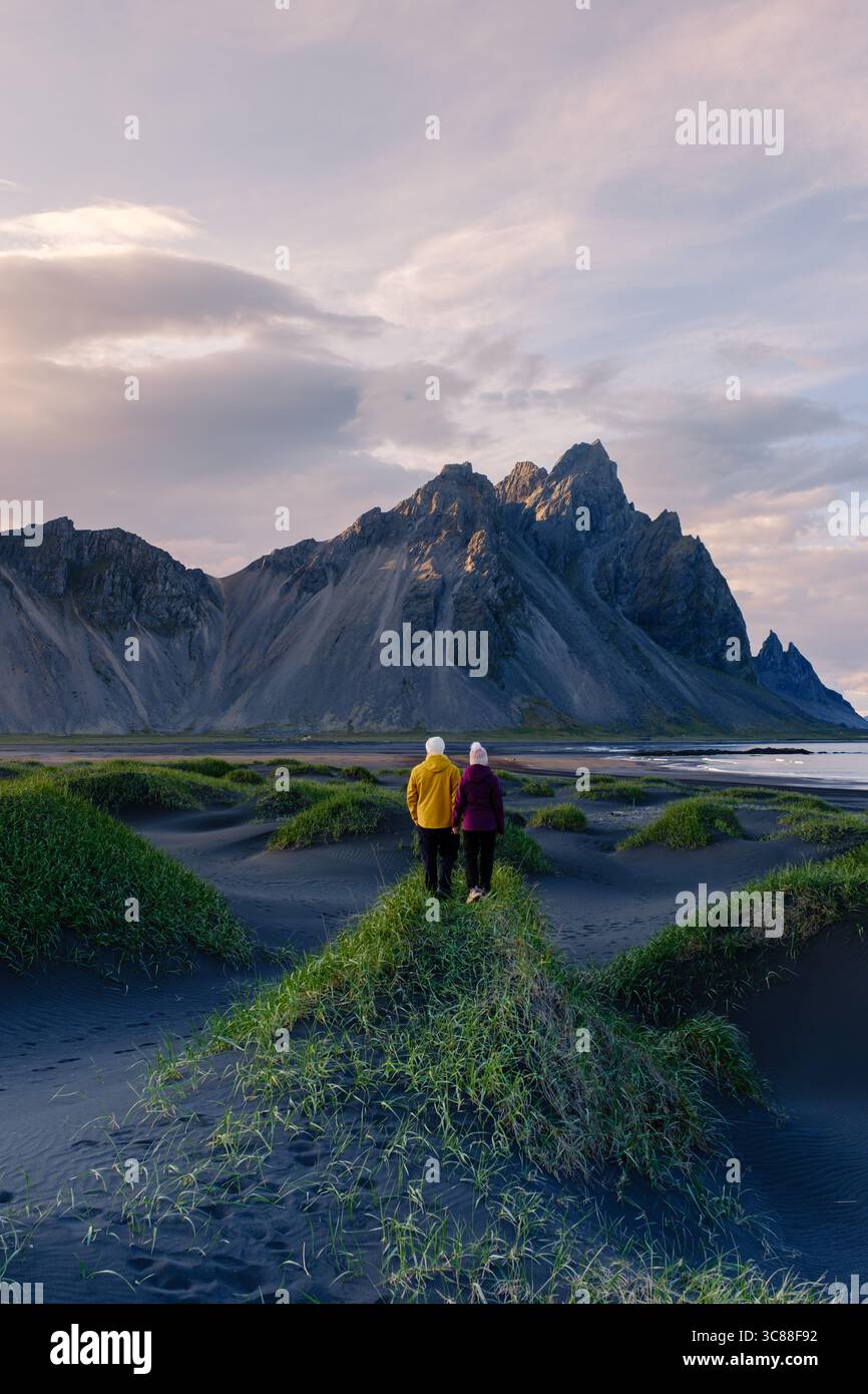 Ein Paar geht Hand in Hand an einem schwarzen Sandstrand Stokksnes Beach Vestrahorn Berg Island, mit beeindruckenden Bergen, die majestätisch im Hintergrund während der goldenen Stunde thronen. Stockfoto