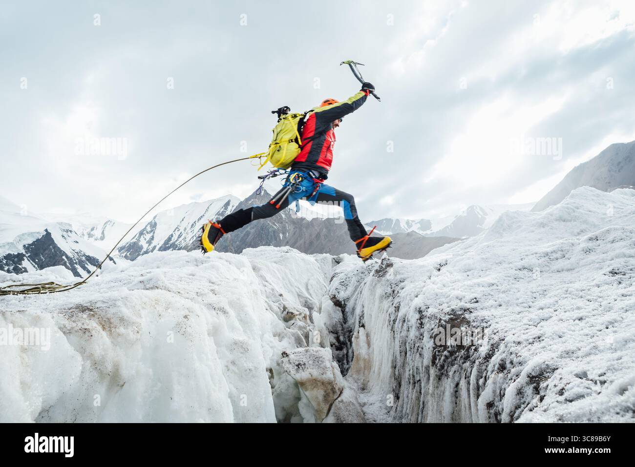 Bergsteiger in pulsierender Ausrüstung hüpft über Gletscherspalten beim Aufstieg des Lenin Gipfels unter klarem Himmel mit schneebedeckten Gipfeln im Hintergrund. Extremer Akt Stockfoto