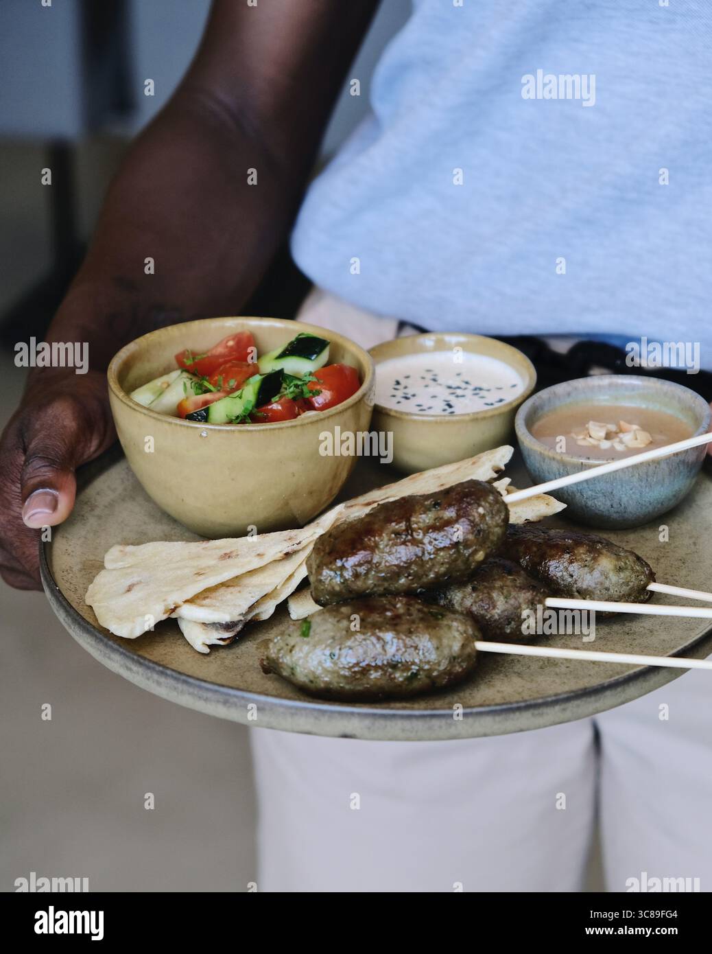 Person mit einer rustikalen Keramikplatte und einer mediterranen Mahlzeit, einschließlich gegrilltem Kofta-Spieß Fladenbrot, Saucen und Salat Stockfoto