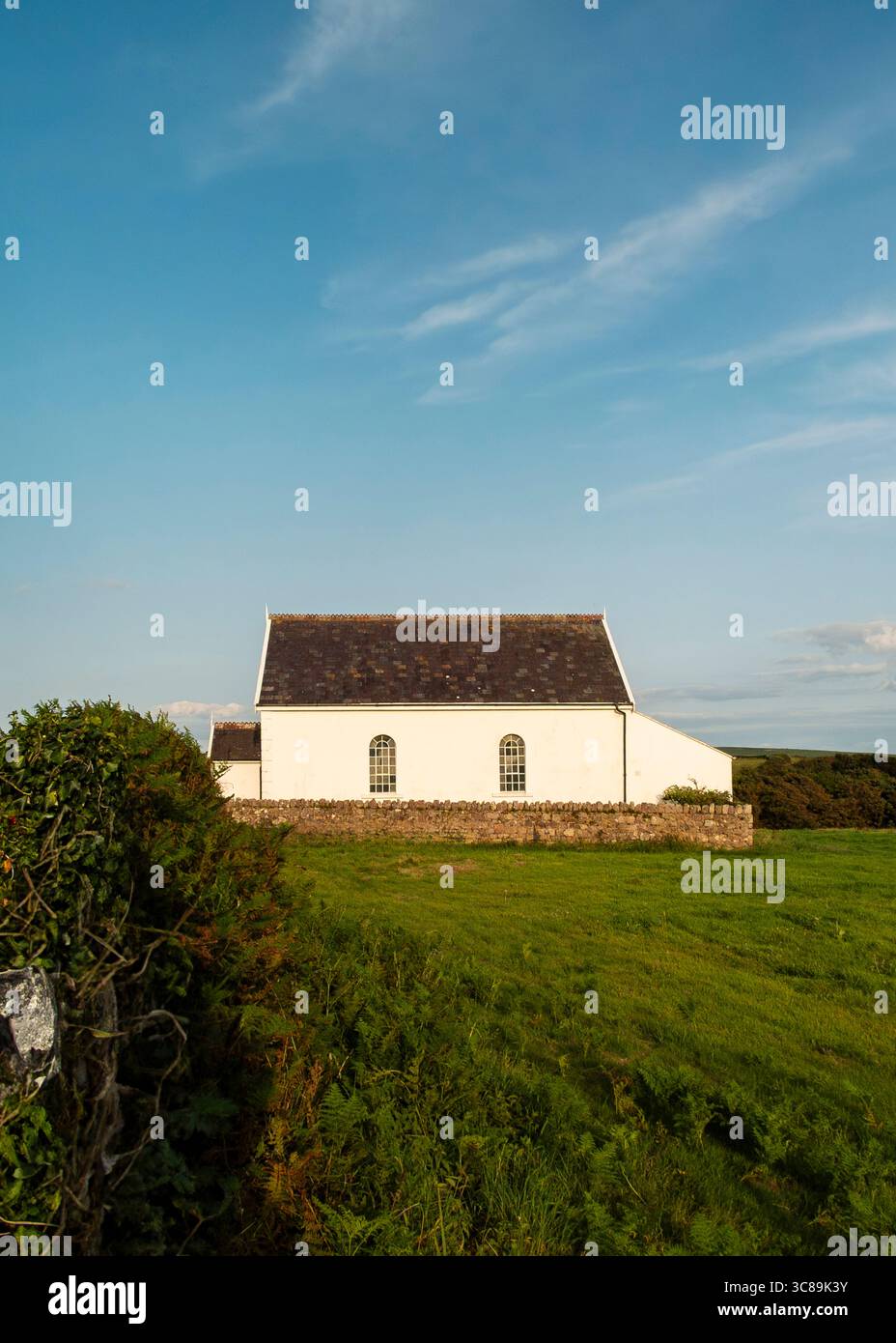 Ein weißes Gebäude mit dunklem Dach befindet sich auf einem grasbewachsenen Hügel mit einer Steinmauer davor. Der Himmel ist blau mit ein paar Wolken. Stockfoto