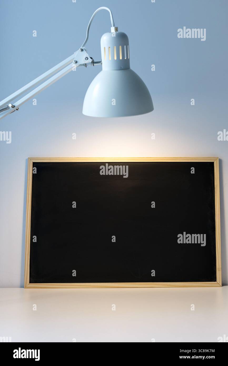 Kreidetafel für Kinder auf dem Tisch, beleuchtet durch eine Lampe, Platz für Beschriftungen und Werbung. Monochromatischer minimalistischer Hintergrund, Zubehör Stockfoto