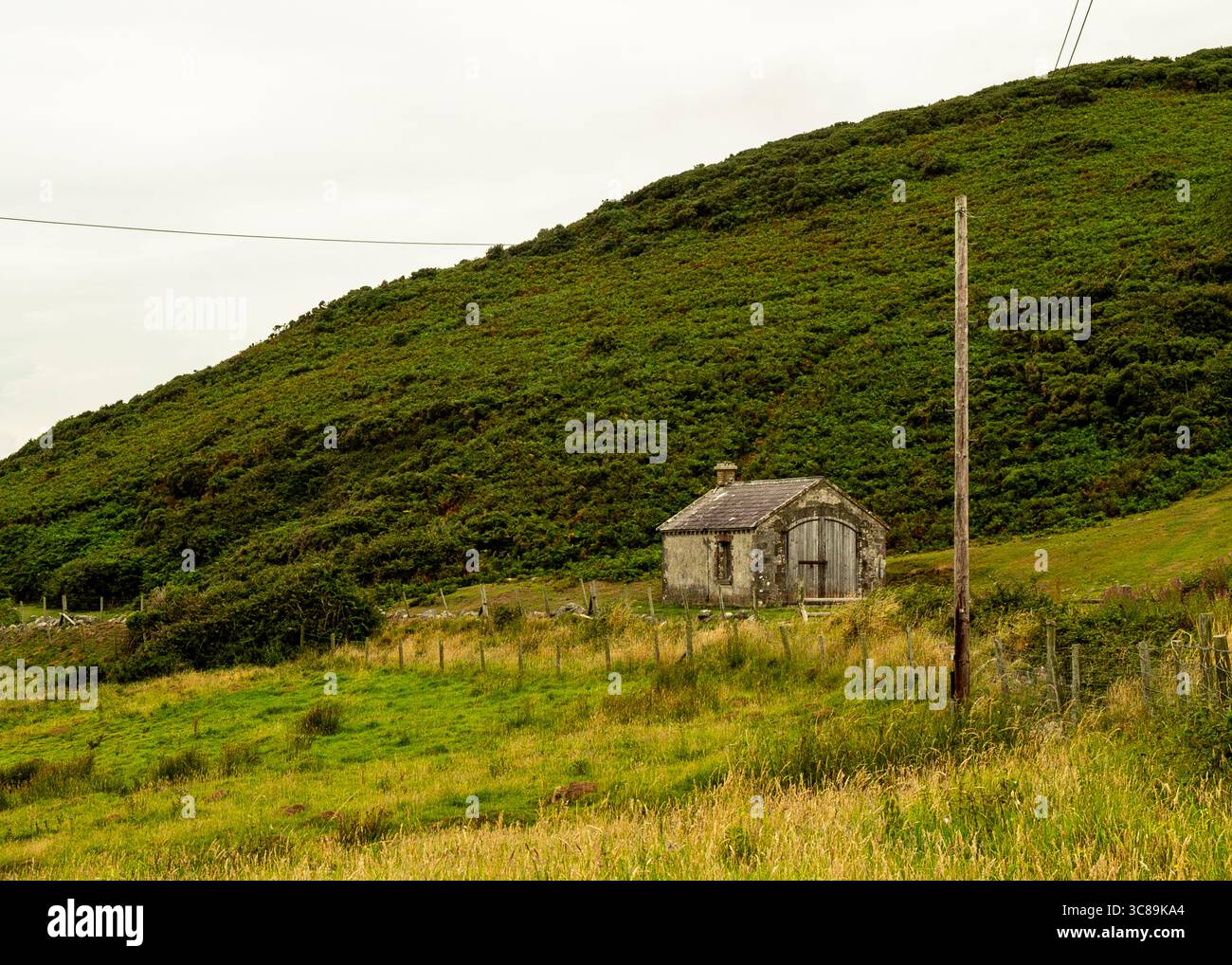 Ein altes Steingebäude befindet sich am Fuße eines großen, grünen Hügels. Ein Holzpfahl steht im Vordergrund, und ein Zaun verläuft am Rand des Feldes entlang. Stockfoto