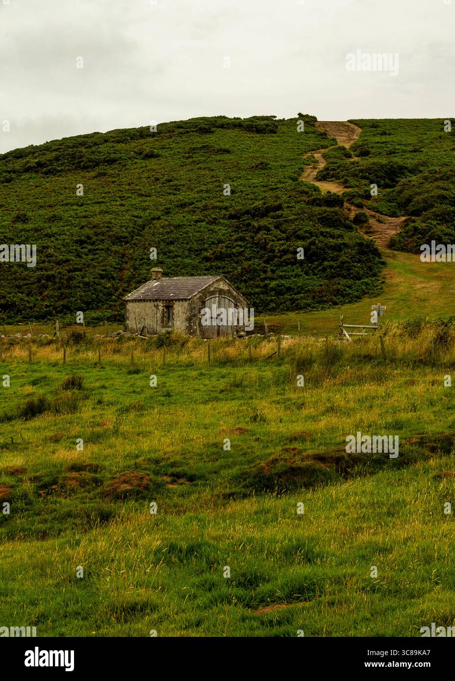 Ein altes Steingebäude befindet sich in einem grünen Grasfeld, mit einem Pfad, der im Hintergrund einen Hügel hinauf führt. Stockfoto