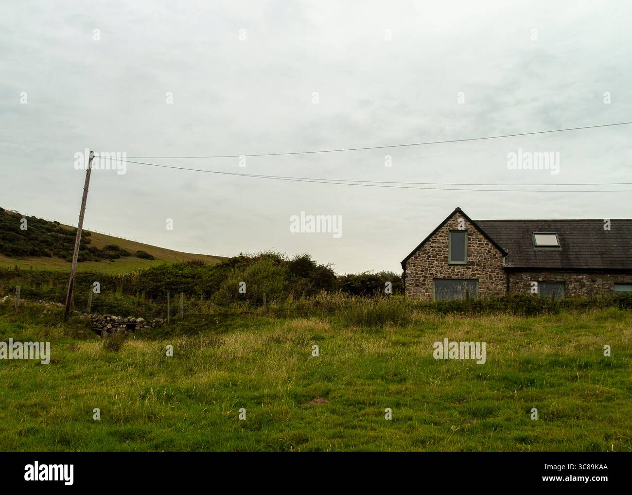 Ein Steinhaus sitzt auf einem Feld aus hohem Gras unter einem bewölkten Himmel. Von der linken Seite des Rahmens erstreckt sich ein Holzmast mit Drähten. Stockfoto