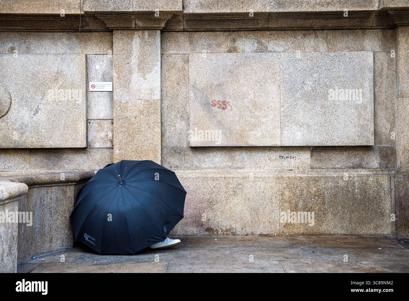 Schwarzer Regenschirm vor verwitterter Steinmauer mit einem Fuß in einer urbanen Straßenszene Stockfoto