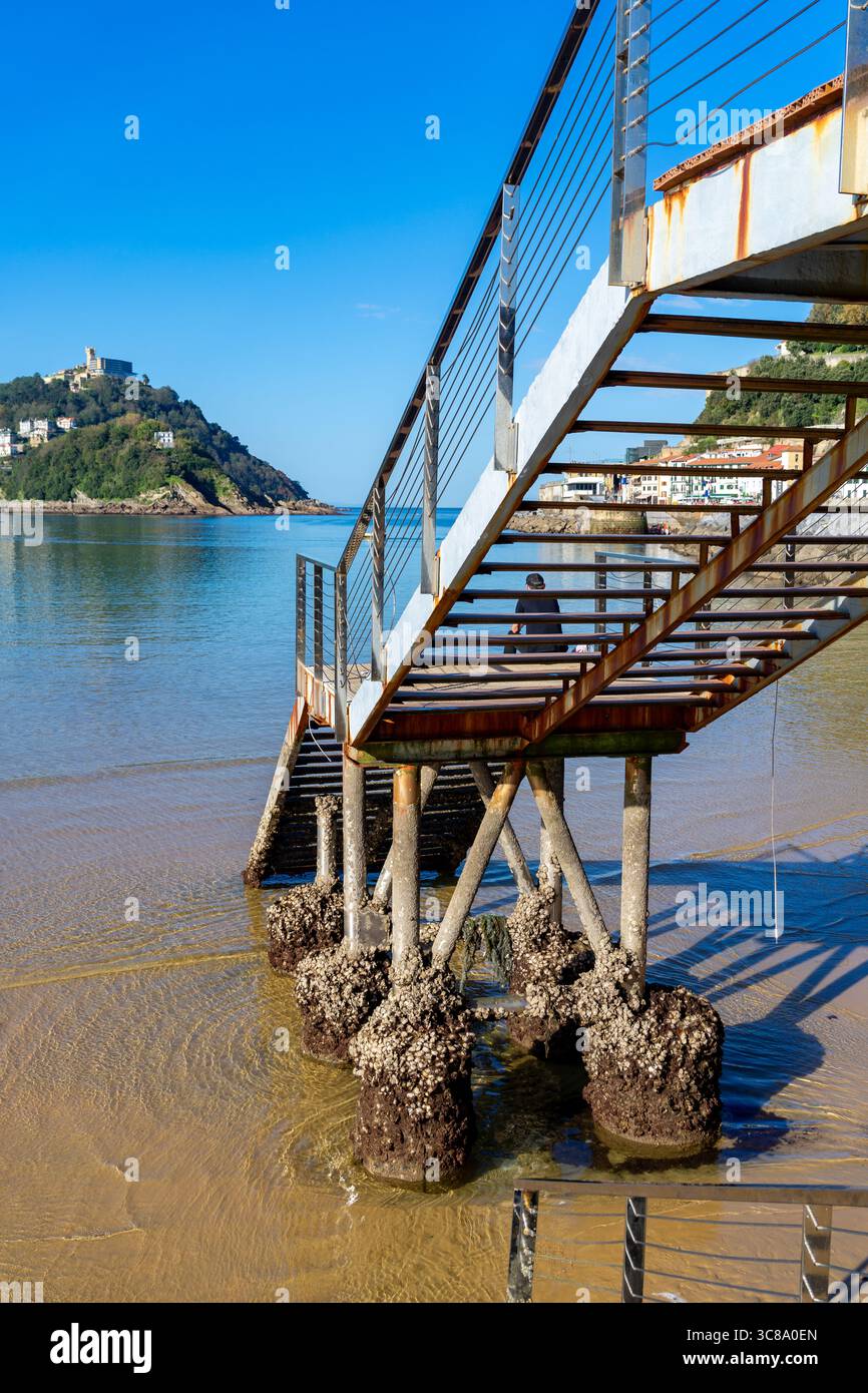 Die Metalltreppen führen hinunter zum Strand und zum Wasser, und eine Person geht die Treppe in der Ferne hoch. Befindet sich in San Sebastian, Spanien. Stockfoto Die Metalltreppen führen hinunter zum Strand und zum Wasser, und eine Person geht die Treppe in der Ferne hoch. Befindet sich in San Sebastian, Spanien. Stockfoto