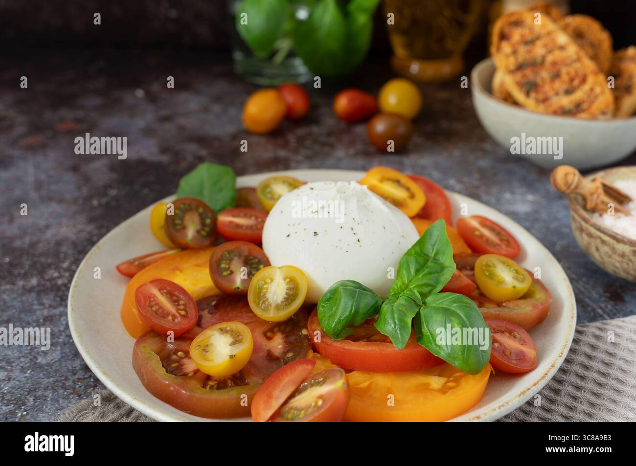 Nahaufnahme von Burrata mit verschiedenen geschnittenen Tomaten, frischem Basilikum und Brot in der Schüssel auf dunkelblaubraunem Hintergrund Stockfoto