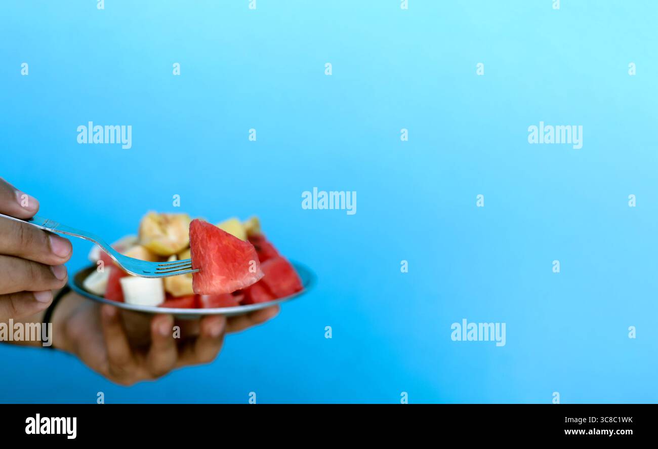 Nahaufnahme einer Hand, die einen Metallteller mit frisch geschnittenen Fruchtstücken wie Banane, Apfel und Wassermelone vor einem leuchtend blauen Hintergrund hält. Ein HE Stockfoto