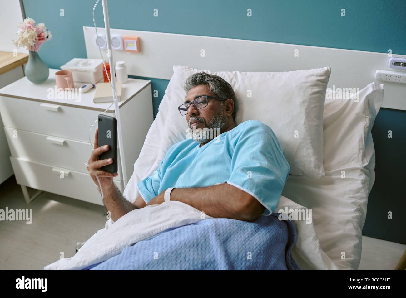 Birassischer Mann, der im Krankenhausbett liegt, mit Smartphone für Kommunikation Stockfoto