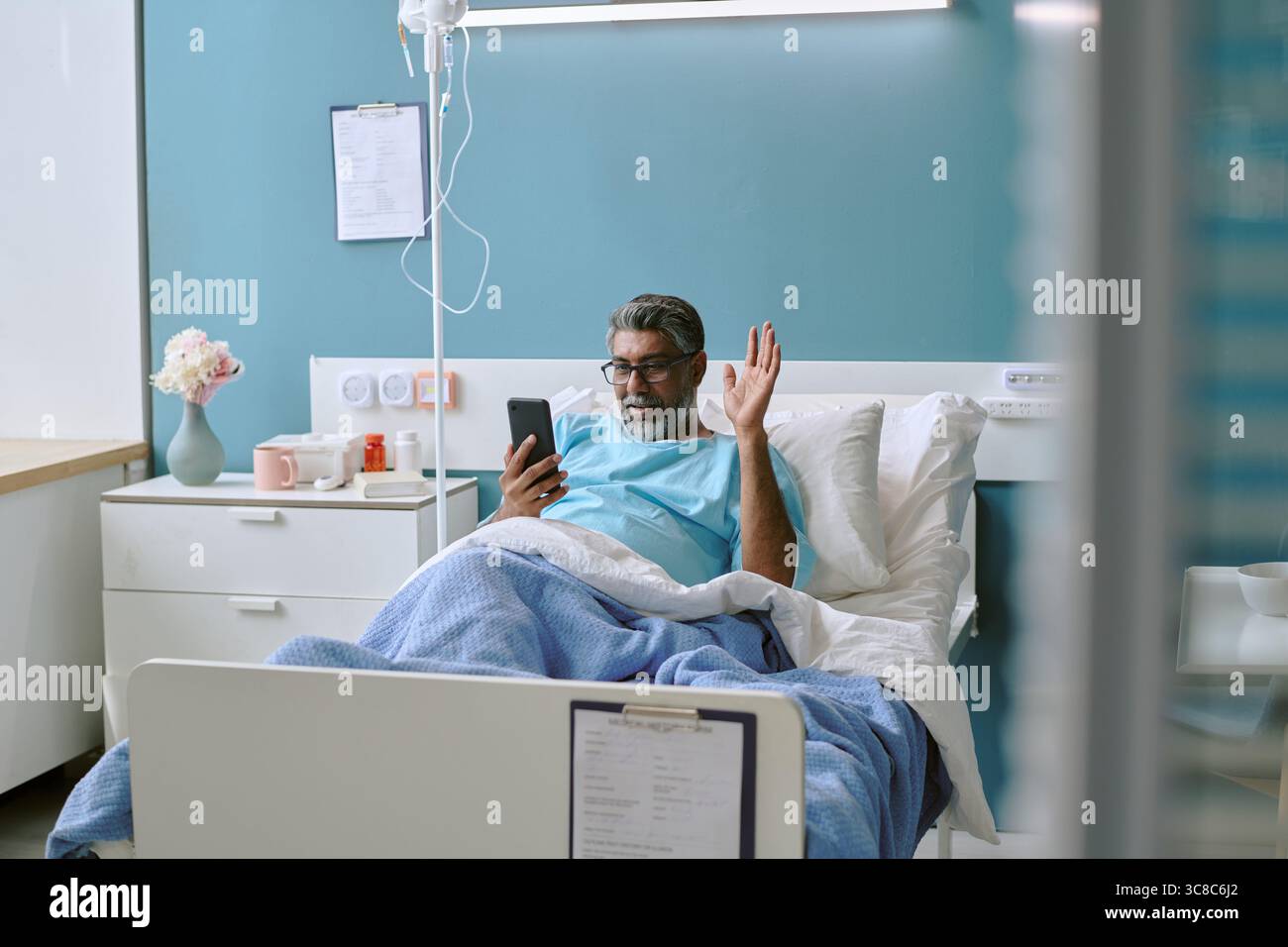 Birassischer Mann mittleren Alters im Krankenhausbett mit Smartphone für Videoanruf Stockfoto