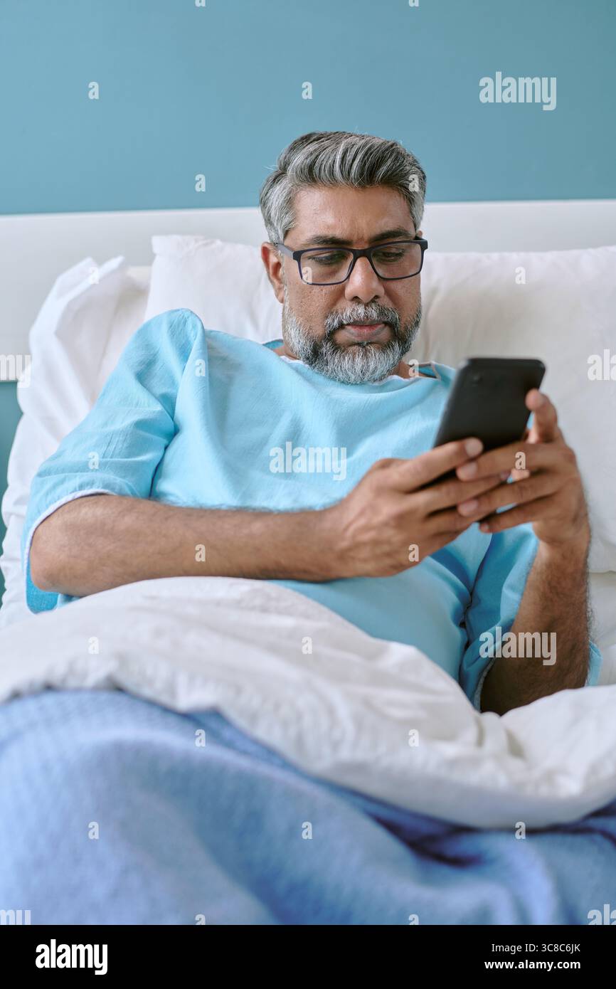 Birassischer Mann mittleren Alters, der mit Smartphone im Krankenhausbett liegt Stockfoto