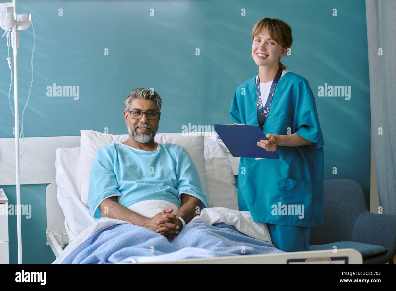 Birassischer Mann mittleren Alters, der im Krankenhausbett sitzt und die junge Erwachsene Krankenschwester lächelt Stockfoto