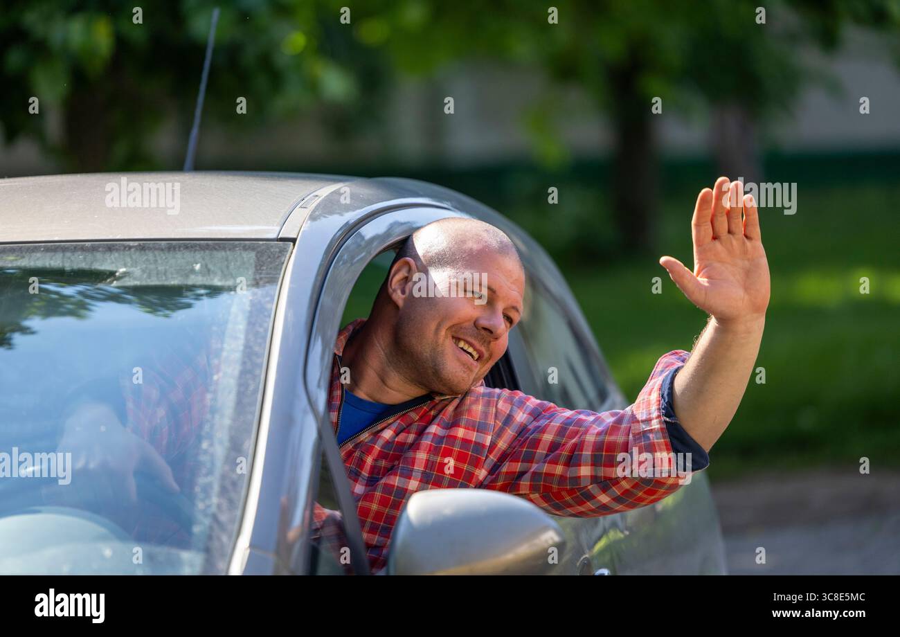 Fröhlicher Mann in kariertem Hemd winkt vom Fahrersitz des geparkten Autos, umgeben von Grün und hellem Sonnenlicht. Stockfoto