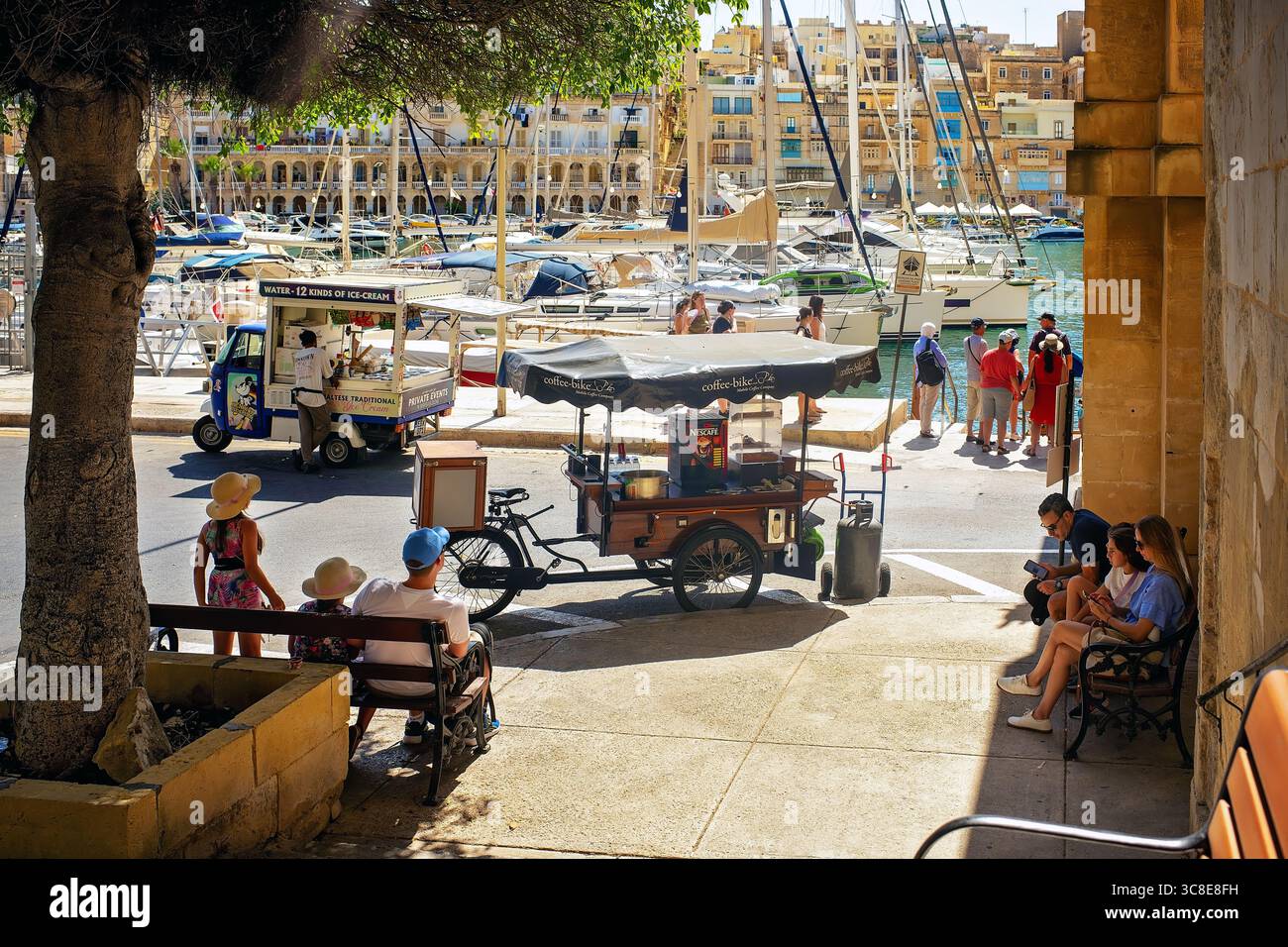 Birgu, Malta – 13. Juli 2025: Touristen genießen eine sonnige Marina Promenade mit lokalen Kaffee- und Eiswagen in der Nähe eines Yachthafens. Stockfoto