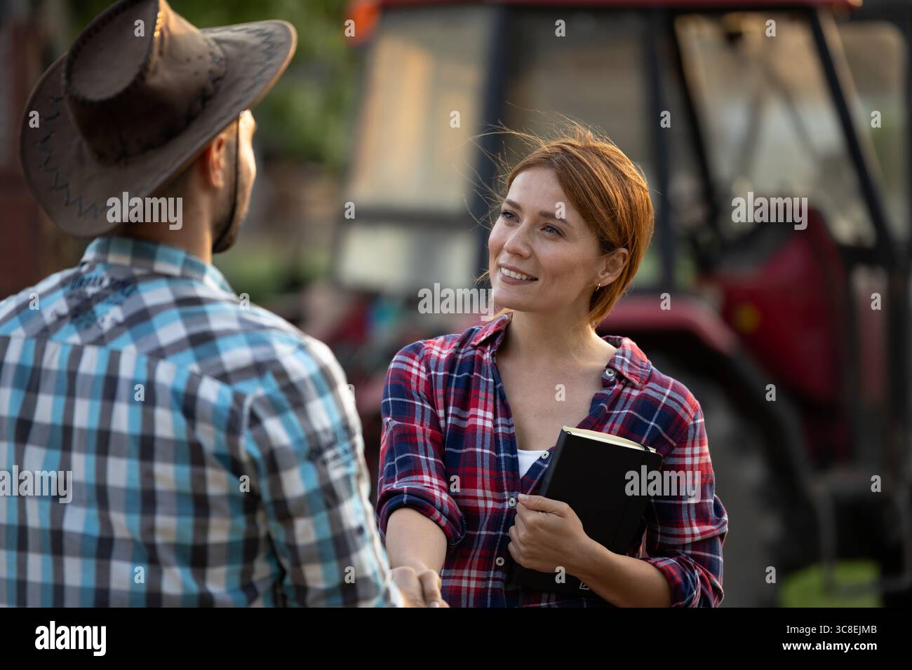 Zwei Bauern, die auf dem Bauernhof handeln, tauschen einen freundlichen Handschlag in der Nähe des Traktors aus. Die Umgebung ist entspannt und einladend, unter einem Abendhimmel Stockfoto