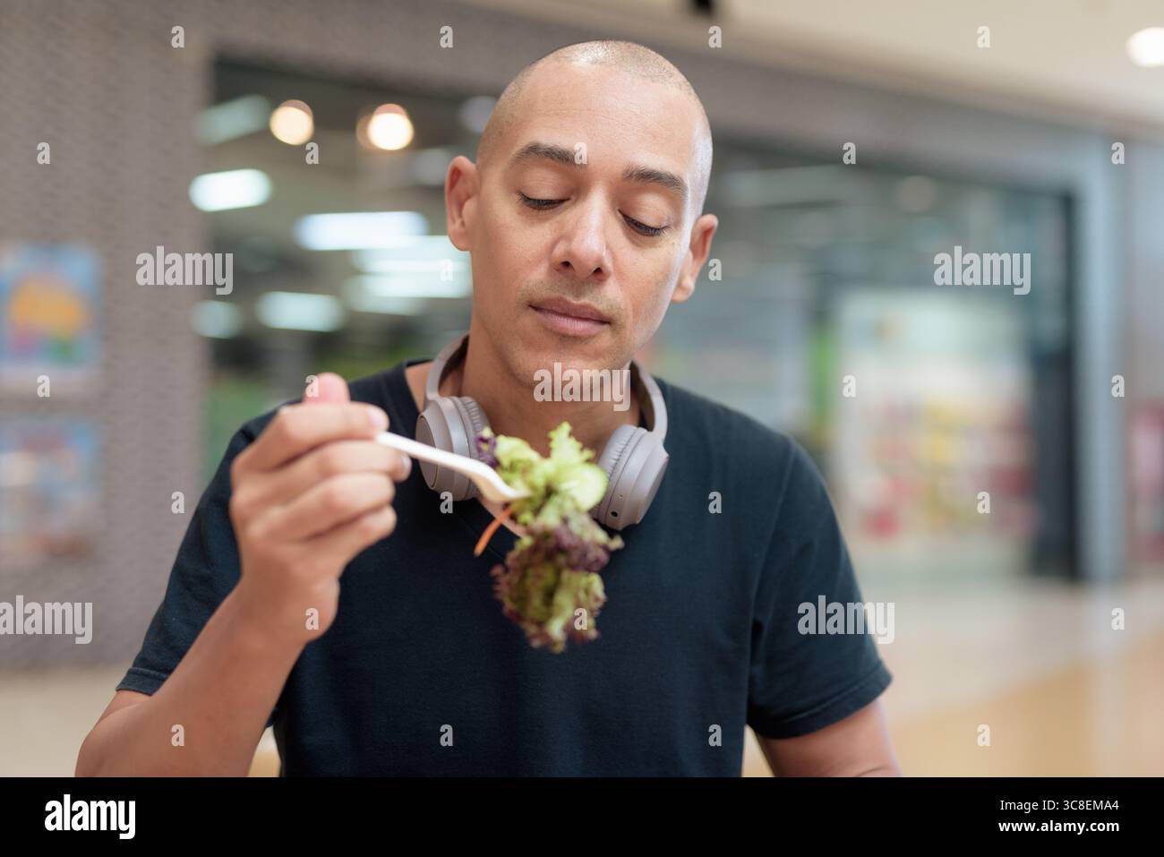 Gemischter kahlköpfiger Erwachsener in schwarzem T-Shirt, der Salat zum Mitnehmen im Einkaufszentrum isst. Gesunder Lebensstil, saubere Ernährung und Ernährung in Real Life setti Stockfoto