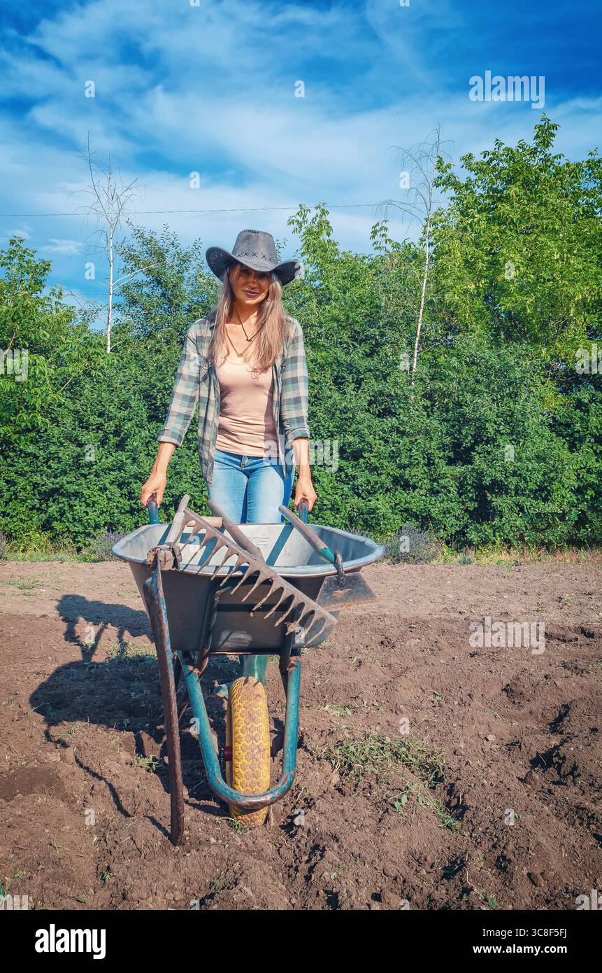 Lächelnde, langhaarige Farmerin in Gummistiefeln mit Schubkarre voller landwirtschaftlicher Werkzeuge gegen Himmel und Bäume. Bio-Gartenbau. Vorderansicht. Stockfoto