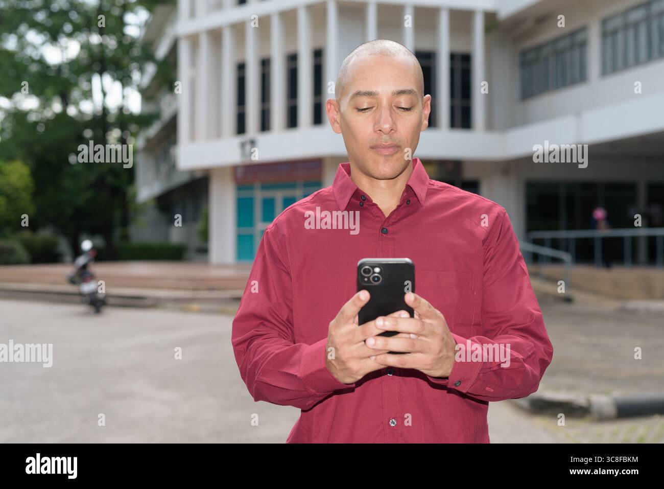 Glatzkopf italienischer Mann in burgunderrotem Hemd, der sein Smartphone überprüft, während er vor dem Universitätsgebäude bei hellem Sonnenlicht steht. Das Konzept des digitalen Kombi Stockfoto