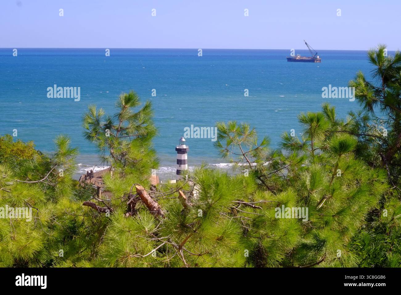 Küstenlandschaft mit Kiefernzweigen, die den Leuchtturm umrahmen, und Frachtschiff, das in ruhigem Wasser vor dem Hintergrund des leuchtend blauen Himmels navigiert Stockfoto