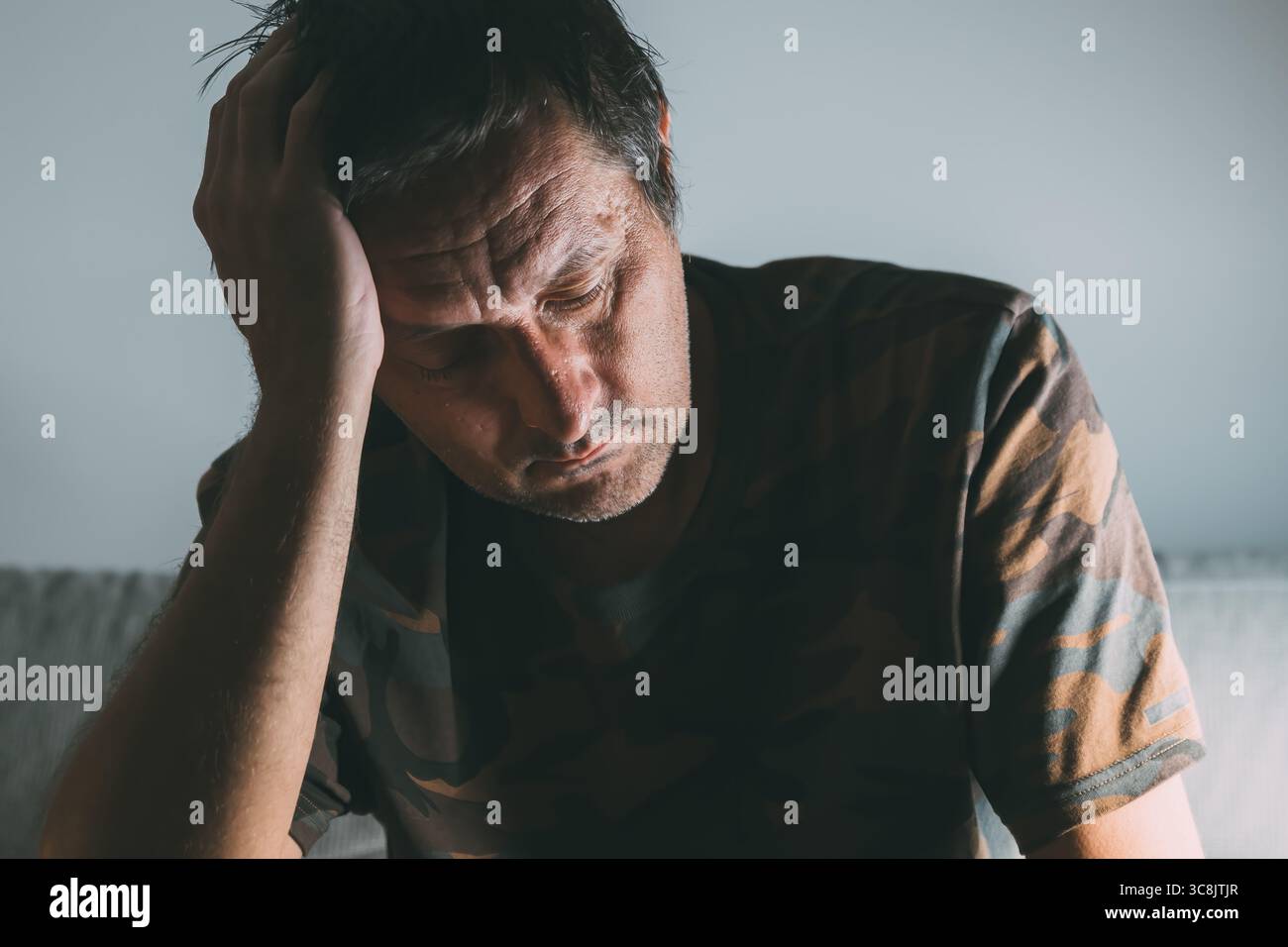Nachdenklicher, bedauernswerter Mann mittleren Alters, der drinnen bei schlechten Lichtverhältnissen sitzt, tief in Gedanken mit Hand auf Kopf, selektiver Fokus Stockfoto